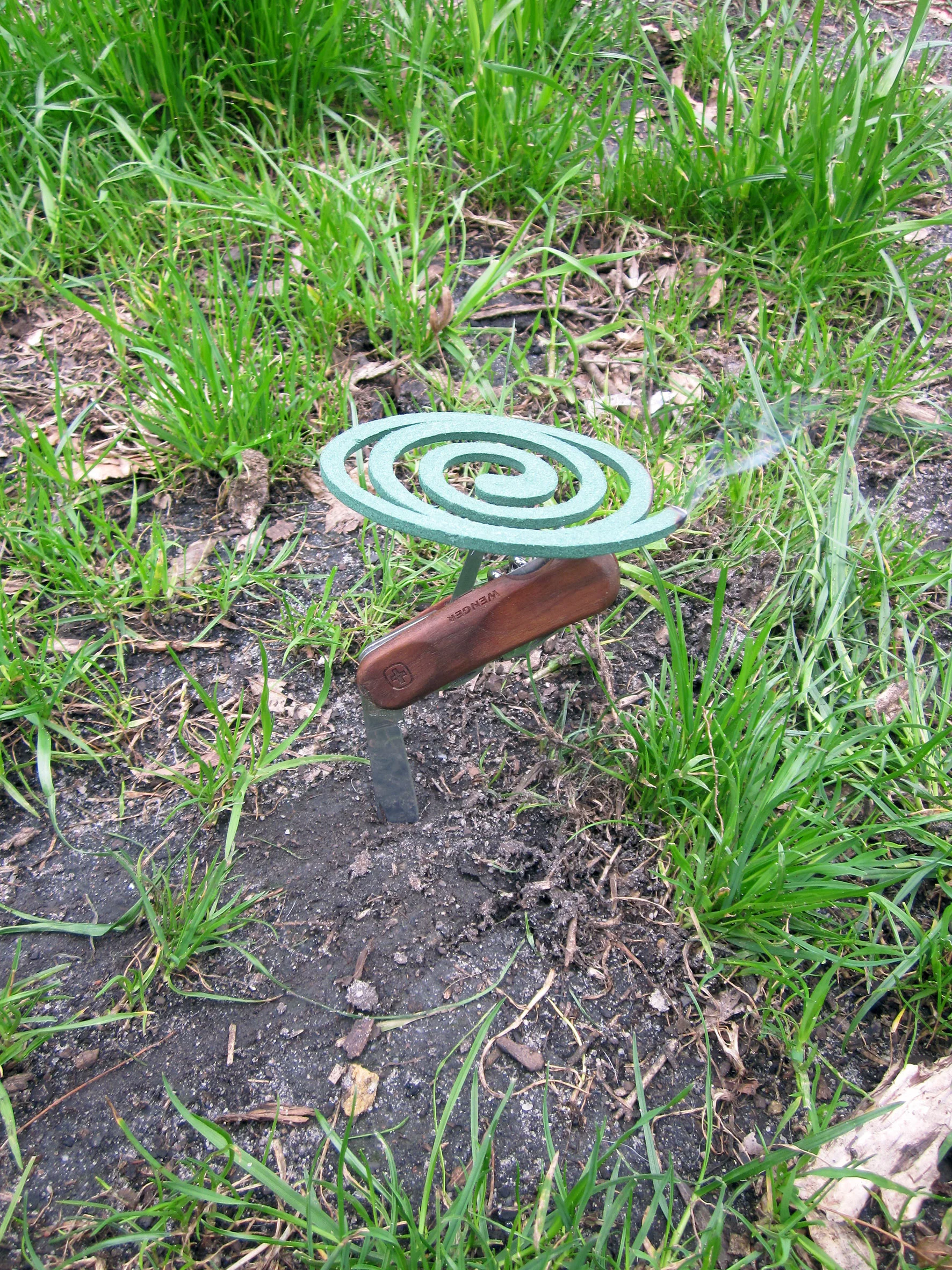 Use any tool on the oppposite side of the main blade on your knife and place the coil on it, the finer point the better. Stab the main blade into the ground and light your coil.