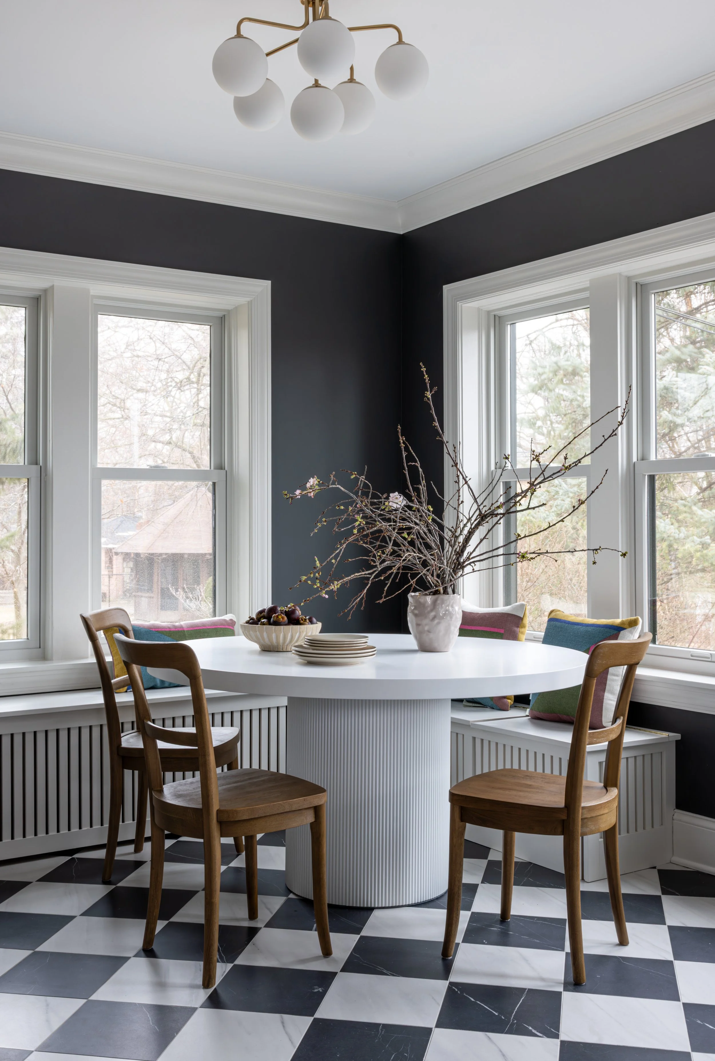 A dining area with a round white table, four wooden chairs, and large windows with a view of trees outside. A white vase with branches and a bowl of fruit are on the table, along with some stacked plates. The room has black walls, white trim, and black-and-white checkered floor tiles.
