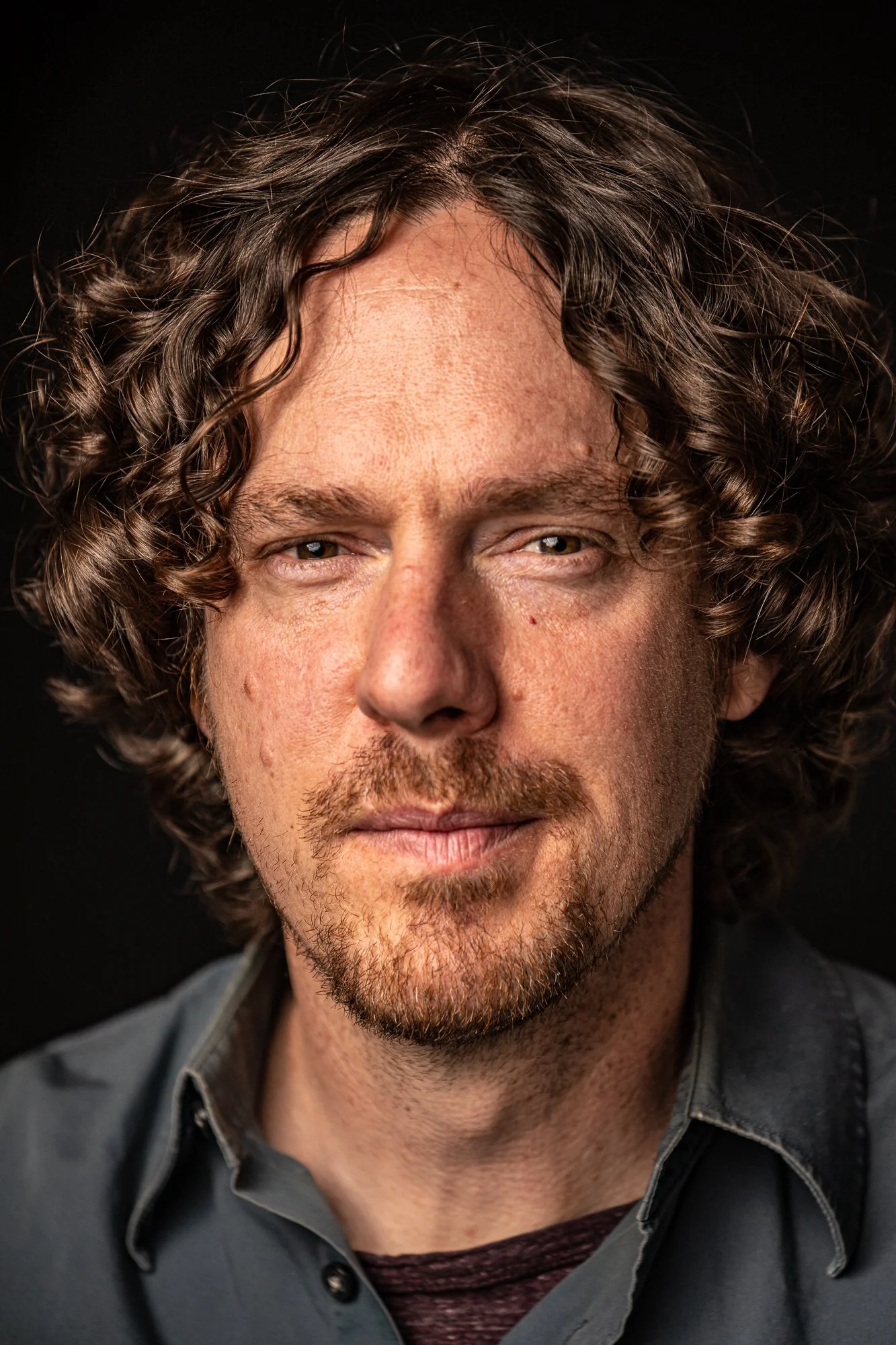 Close-up portrait of a man with curly hair and facial hair, wearing a gray shirt.