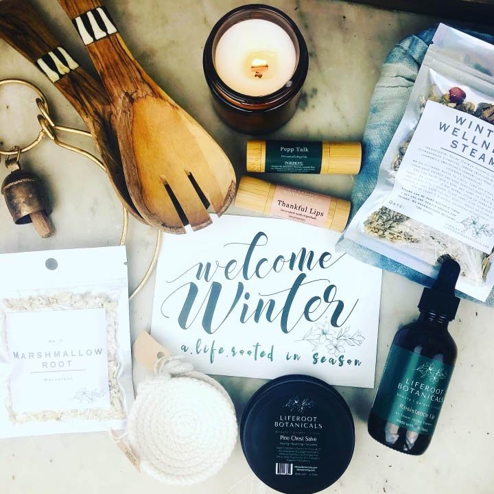 Assortment of winter-themed wellness products including a "Welcome Winter" sign, wooden kitchen utensils, a candle, essential oils, wellness steam packet, lip balm, marshmallow root, and a white crocheted scrubby.