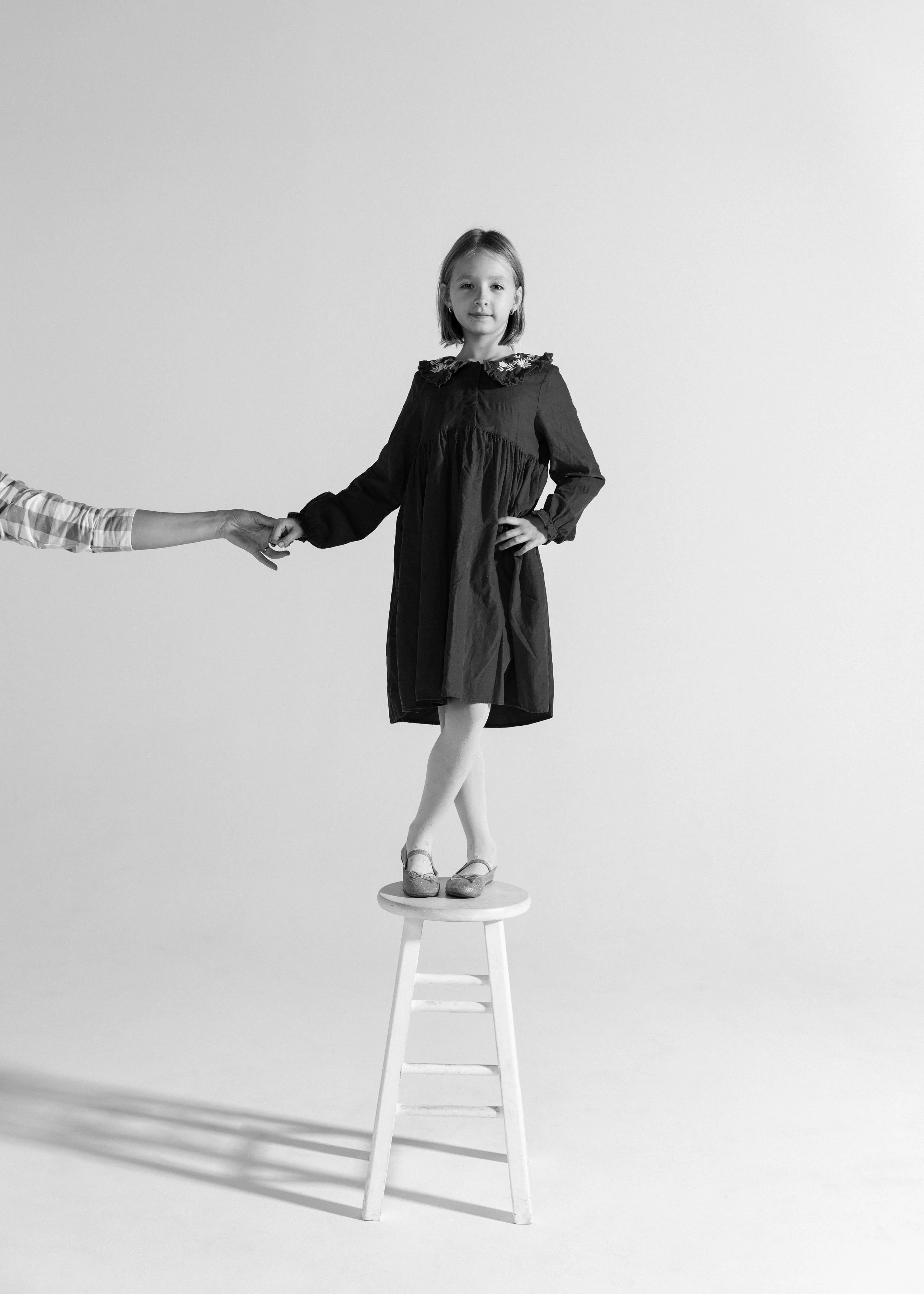 A young girl stands on a stool, holding hands with another person who is partially out of frame. The girl is wearing a dark dress and shoes, and has a confident expression against a plain background.