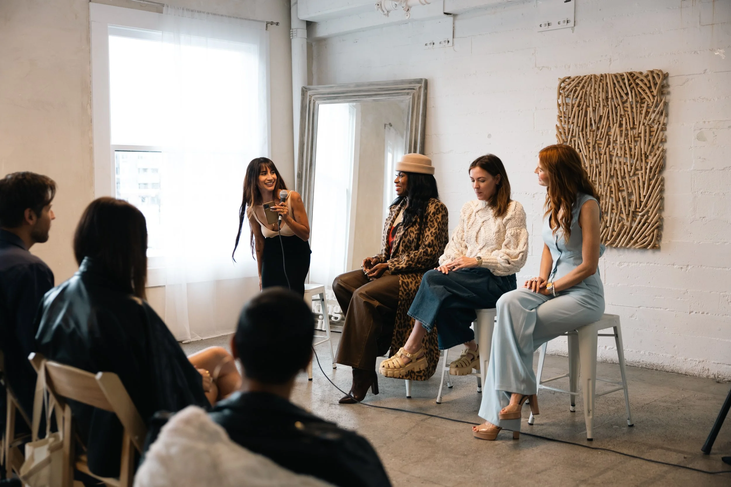 Four women sitting on stools on a stage, one woman standing and speaking into a microphone, in front of an audience, in an indoor venue with natural light.