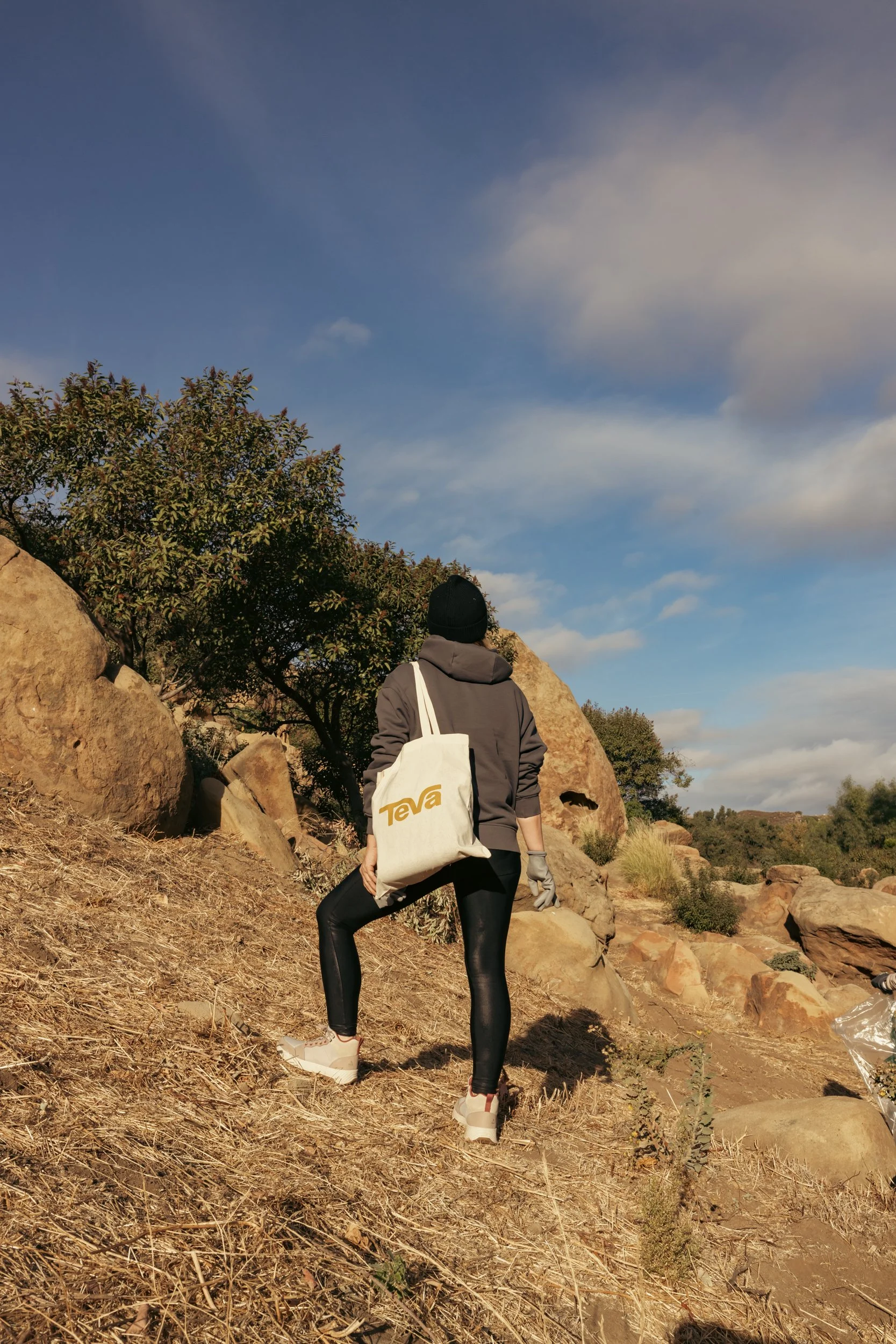 Person hiking outdoors with a backpack in a rocky and desert-like landscape under a partly cloudy sky.