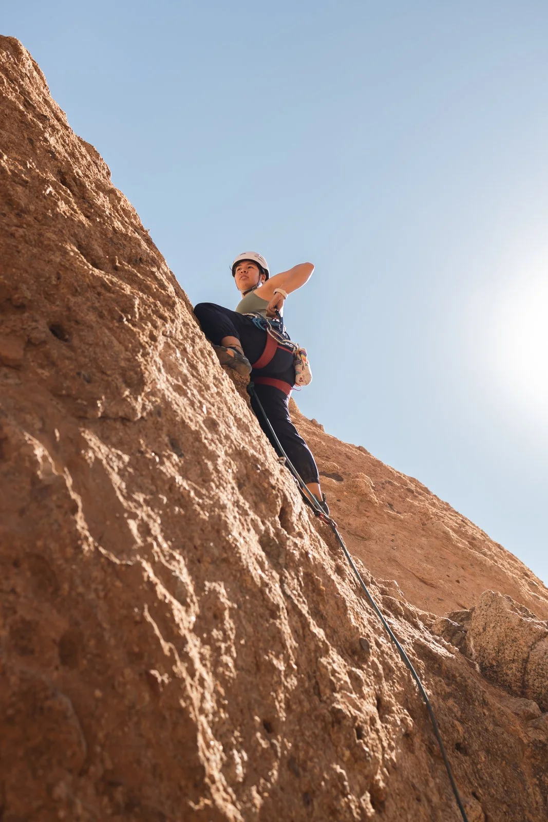 A woman rock climbing on a steep, rugged cliff under a clear blue sky, wearing a safety helmet and climbing gear.