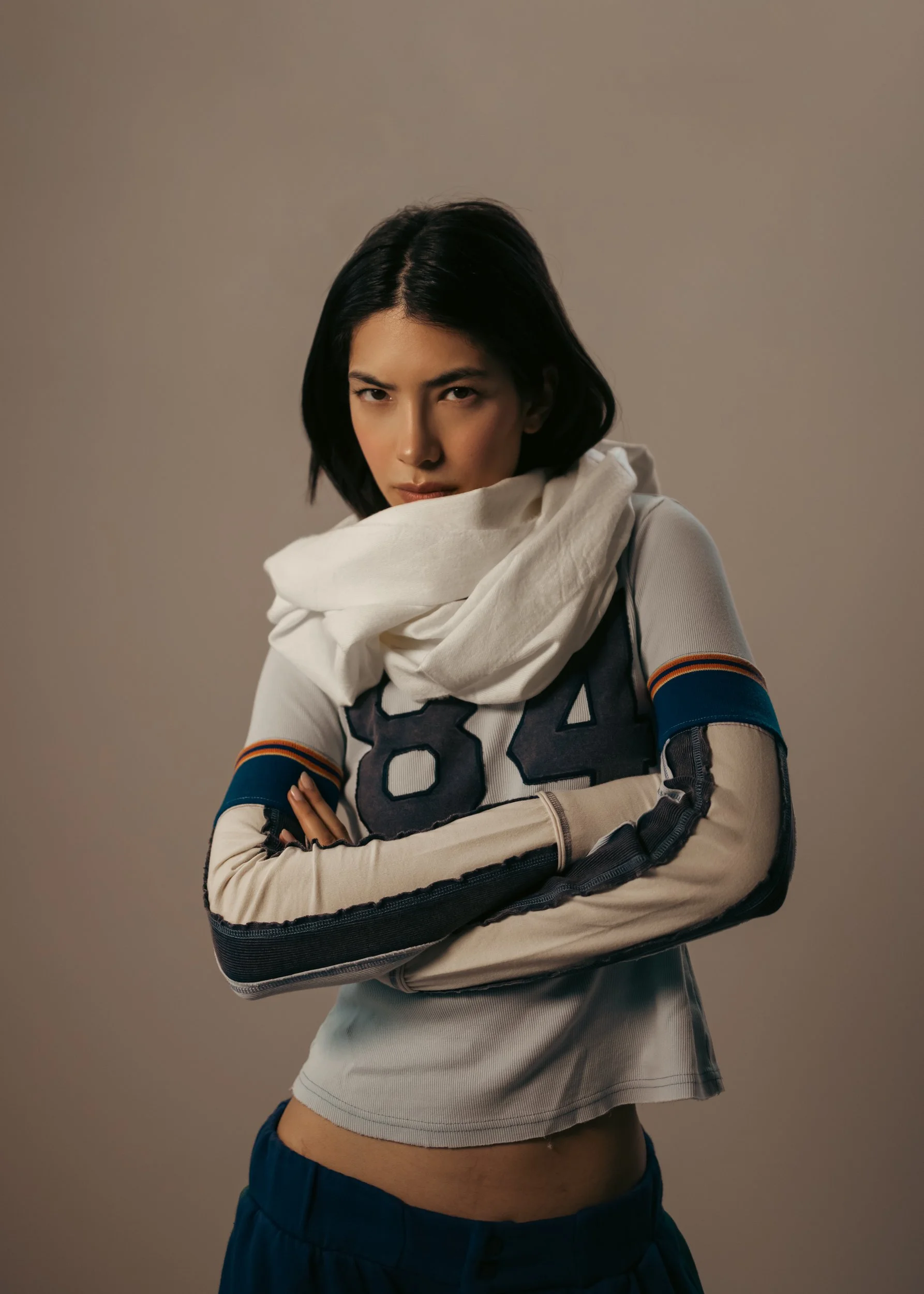 A woman with short black hair wearing a white sports jersey with the number 84, a white scarf, and dark pants, stands with arms crossed against a plain neutral background.
