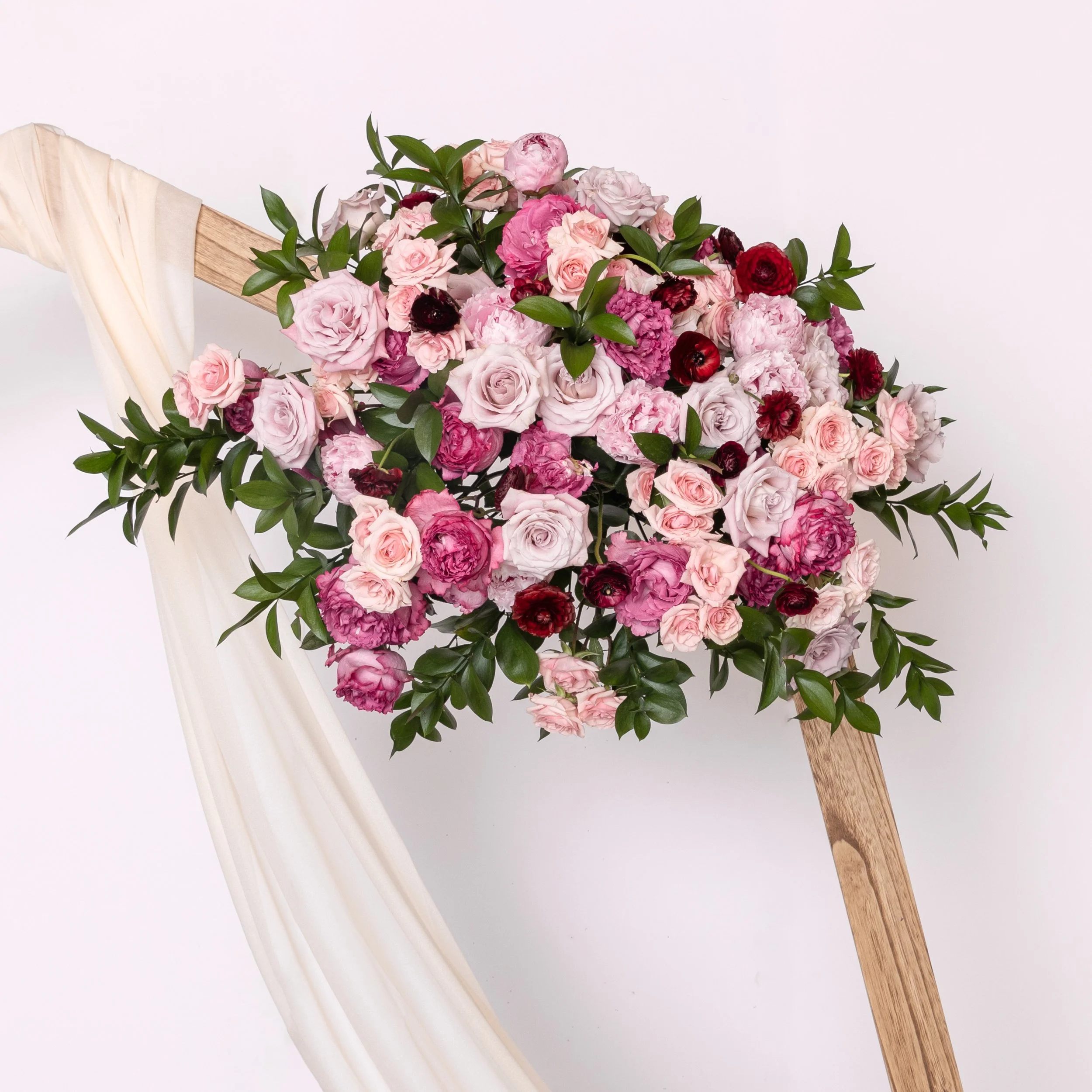Arrangement of pink and burgundy roses and greenery on a wooden stand with white fabric draped over it.