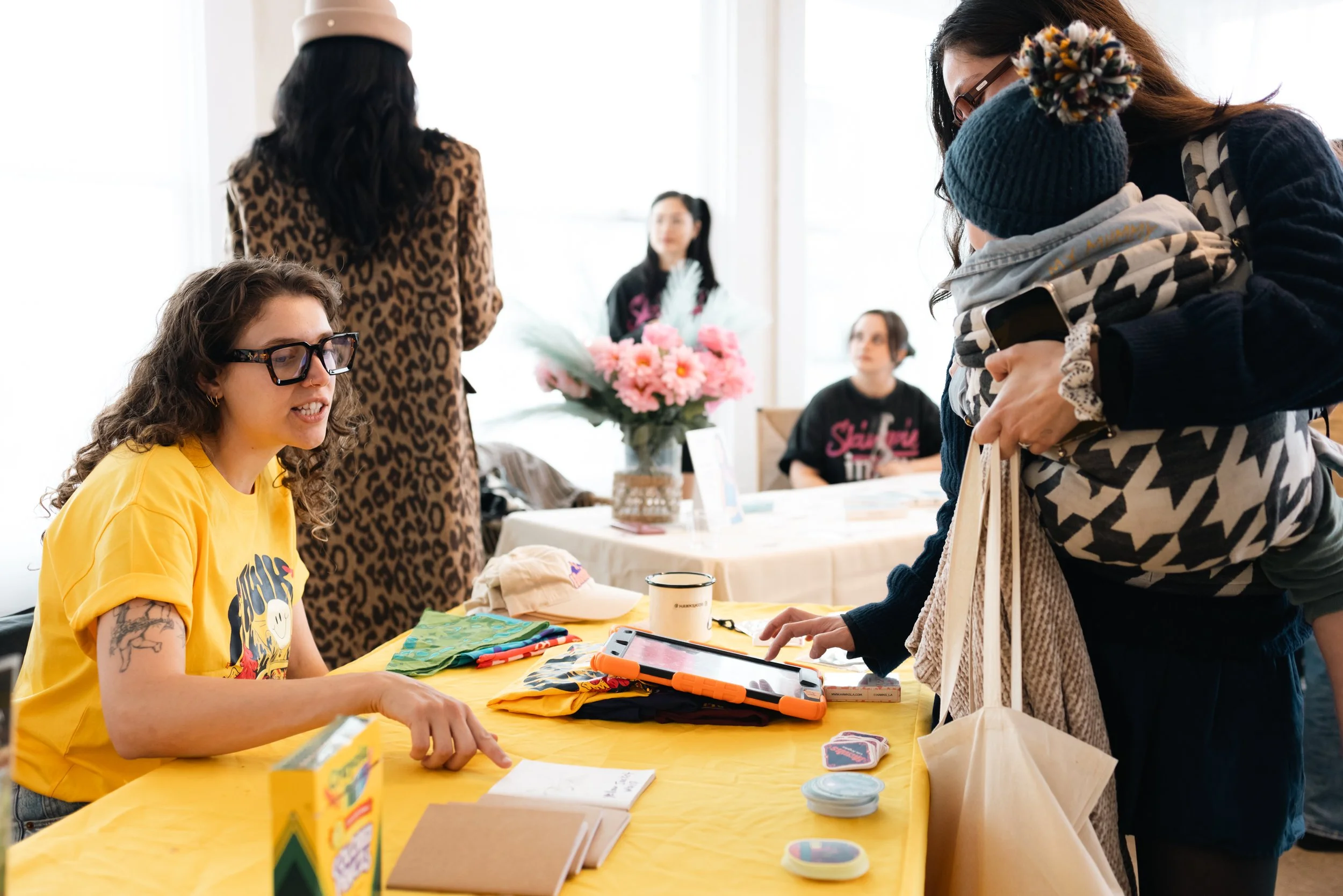A woman with glasses and curly hair is sitting at a table covered with a yellow cloth, talking to a woman standing across from her holding a child. The table has colorful pens, a notebook, a cup, and small items. Behind them, another woman with pigta
