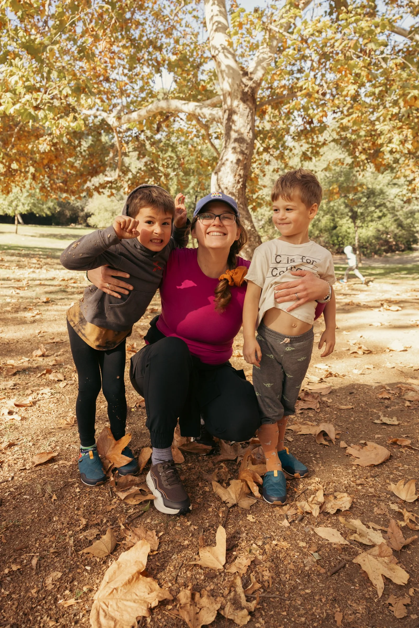 A woman and two young boys smiling and posing outdoors on a fall day with autumn leaves on the ground and a large tree in the background. Outdoor family event