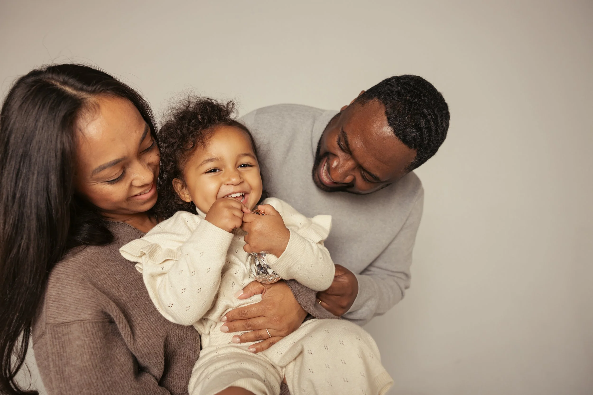 A happy family of three laughing together, with a woman and man holding a young girl, against a plain light-colored background.