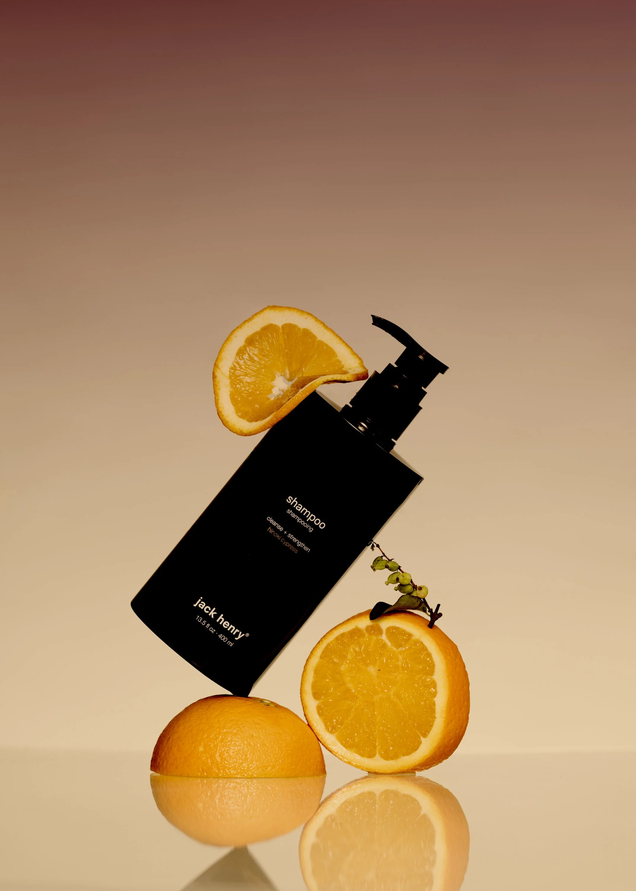 A black shampoo bottle with a slice of orange on top, positioned between two halved oranges, with one orange on a reflective surface.