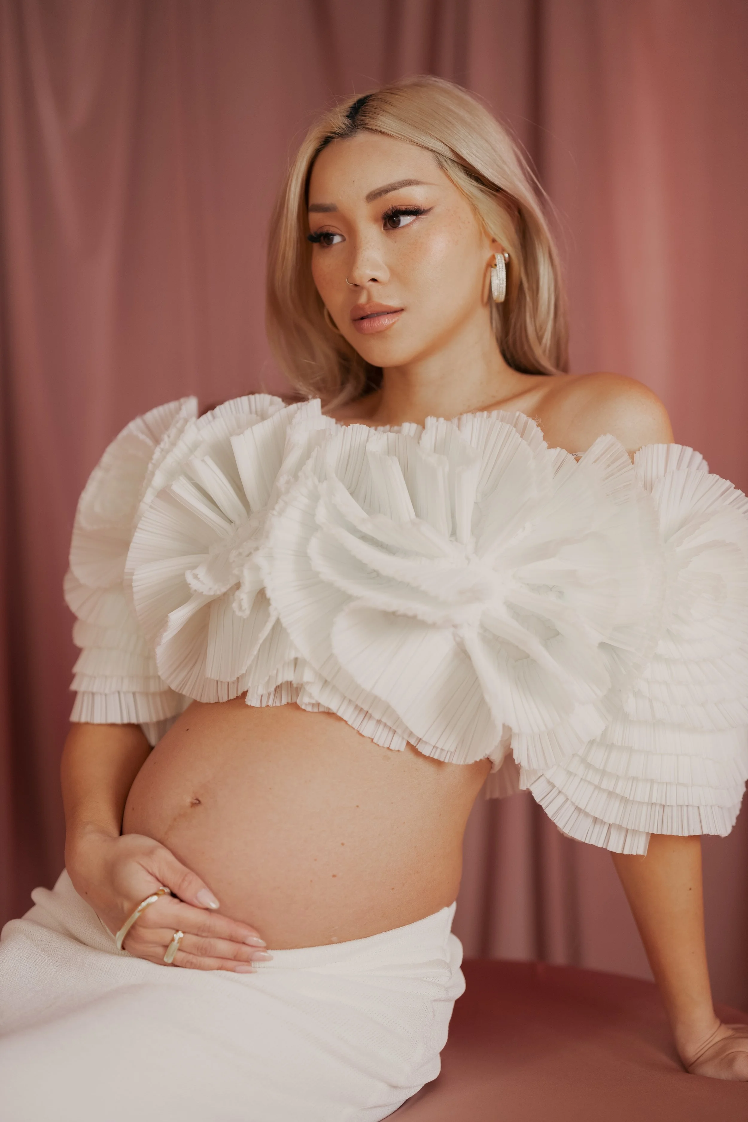 A pregnant woman with blonde hair sitting in front of pink curtains, wearing a white off-the-shoulder ruffled top and a white skirt.