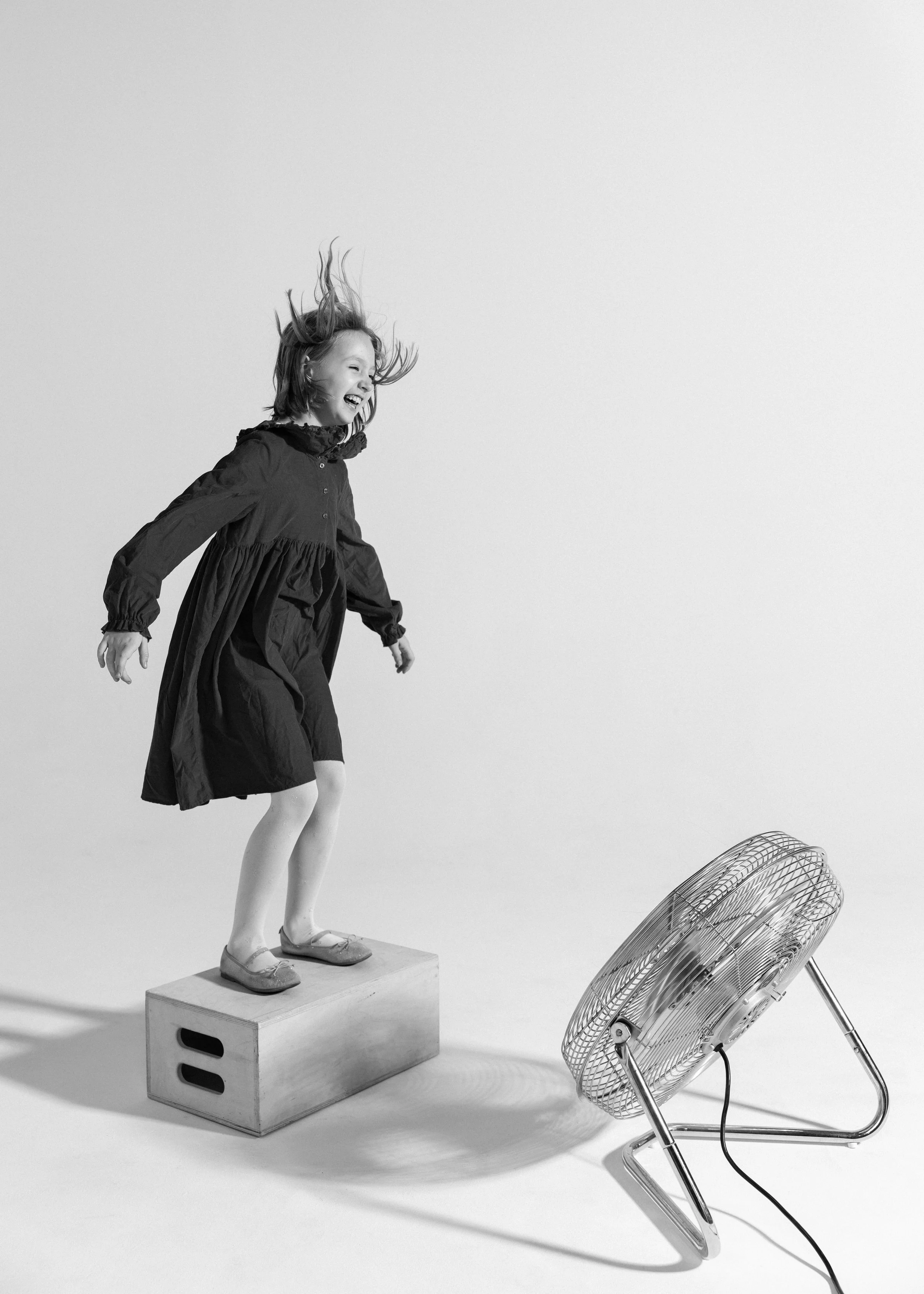 A young girl in a dark dress standing on a wooden block, smiling as she faces a large electric fan and wind blows her hair.