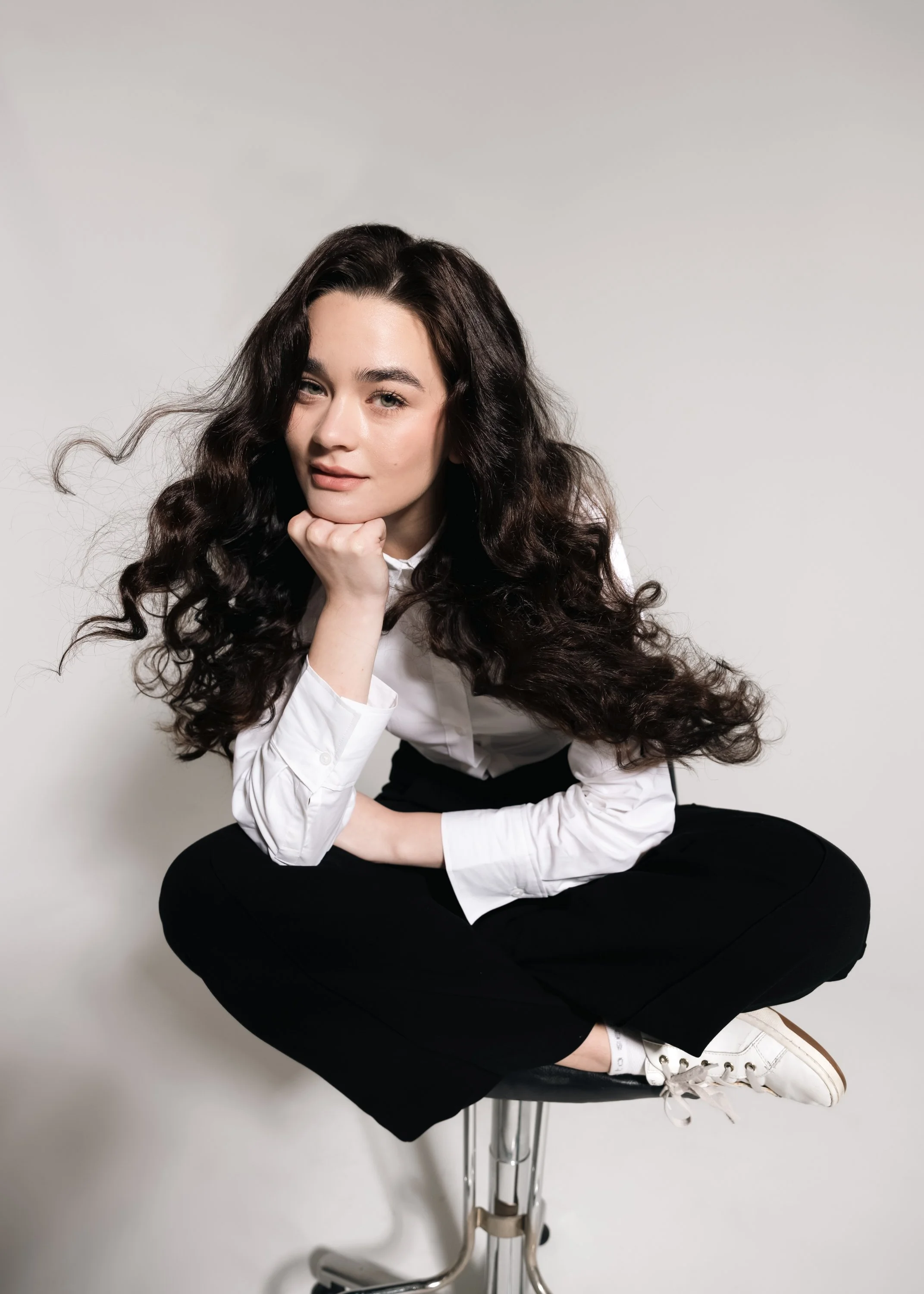 A young woman with long, wavy dark hair, wearing a white shirt, black pants, and white sneakers, sitting cross-legged on a stool against a plain white background.