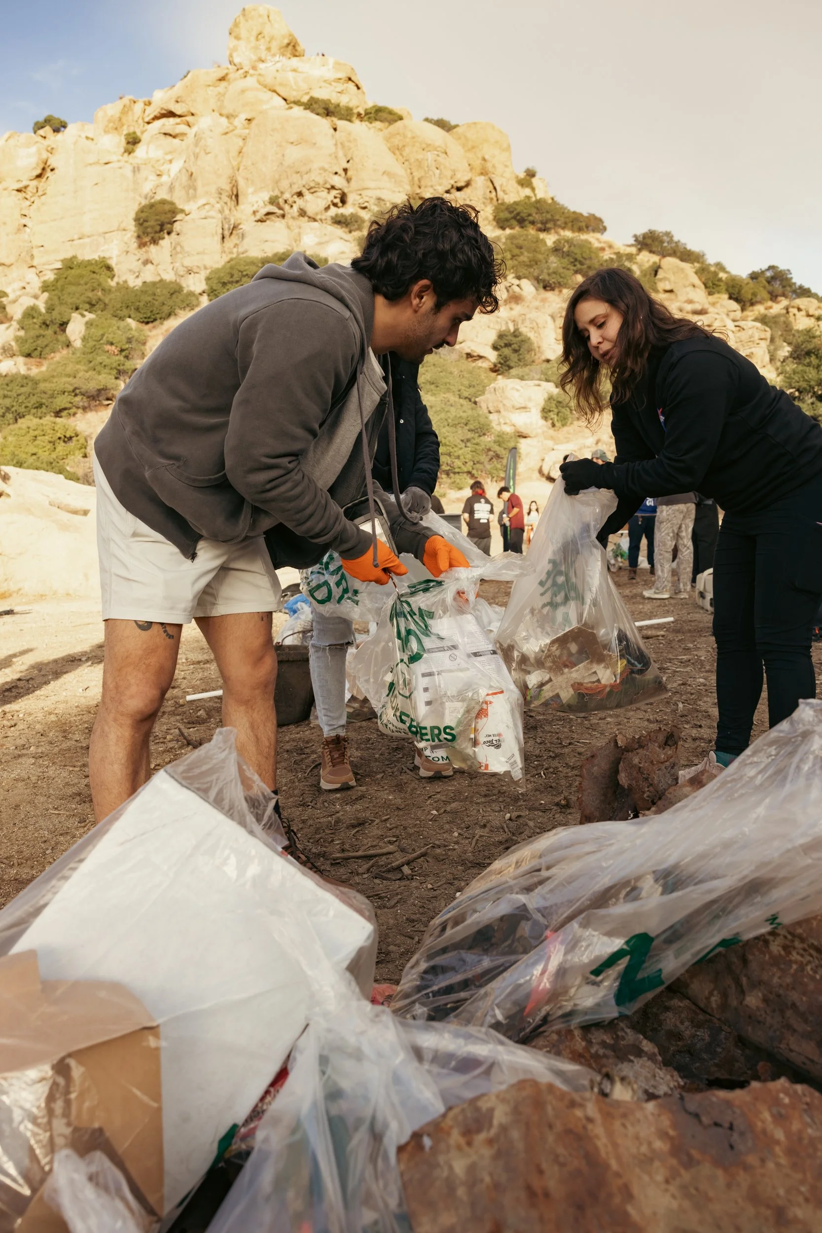 People participating in a cleanup event, collecting trash in a rocky outdoor area with a mountainous background.