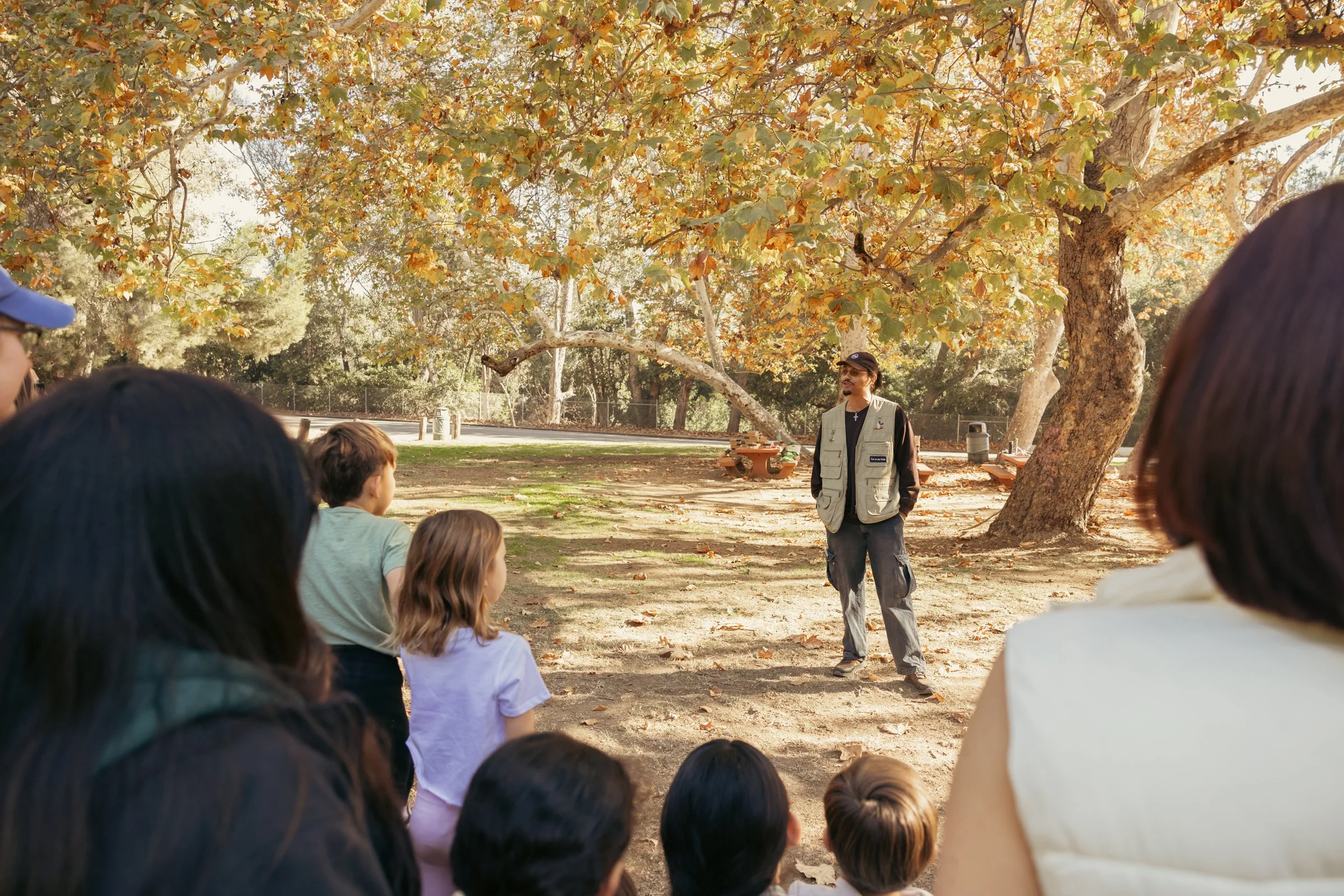 A group of children and adults listening to a park ranger or guide under a large tree with autumn-colored leaves.
