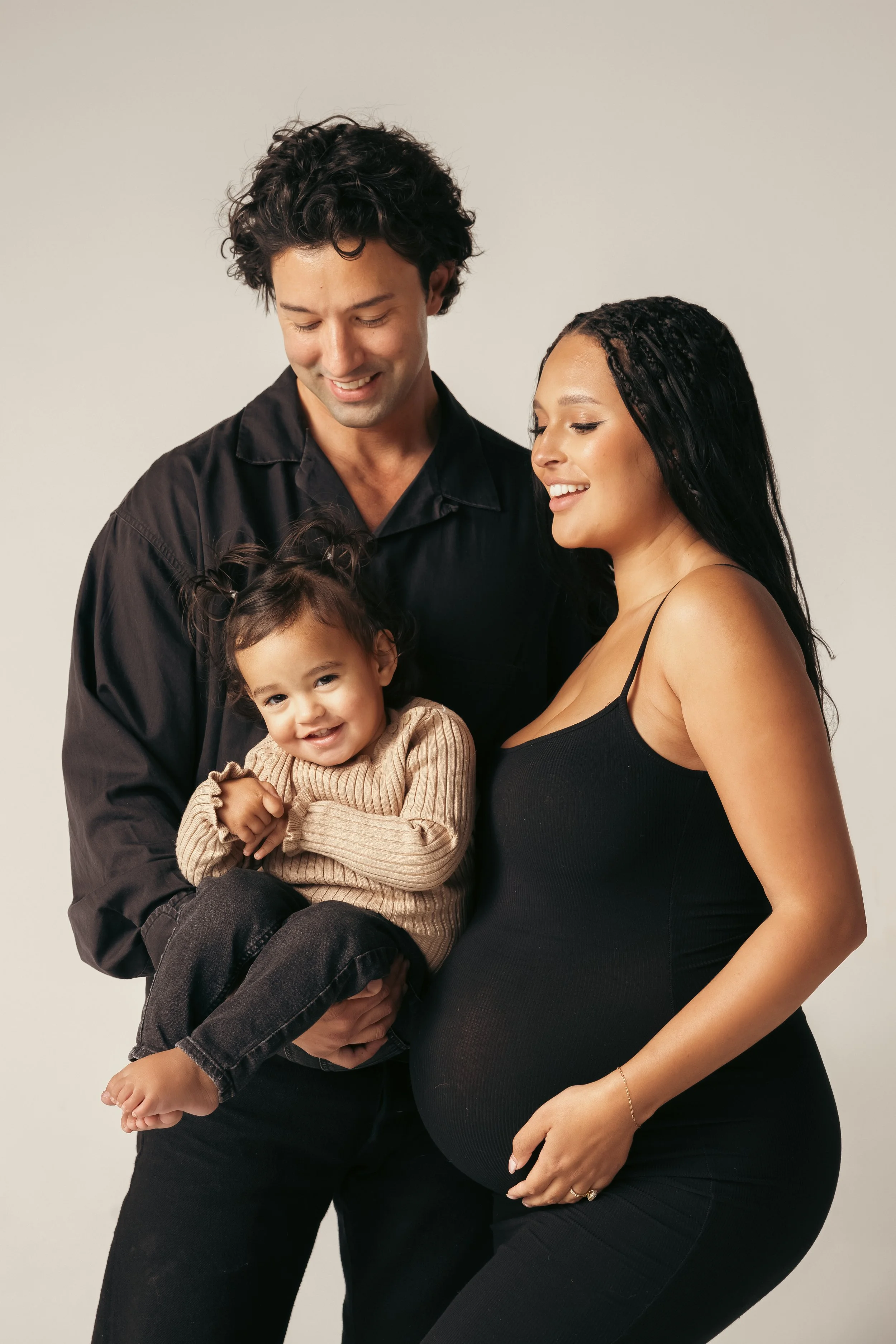 A smiling family with a pregnant woman, man, and young girl standing together against a plain background.
