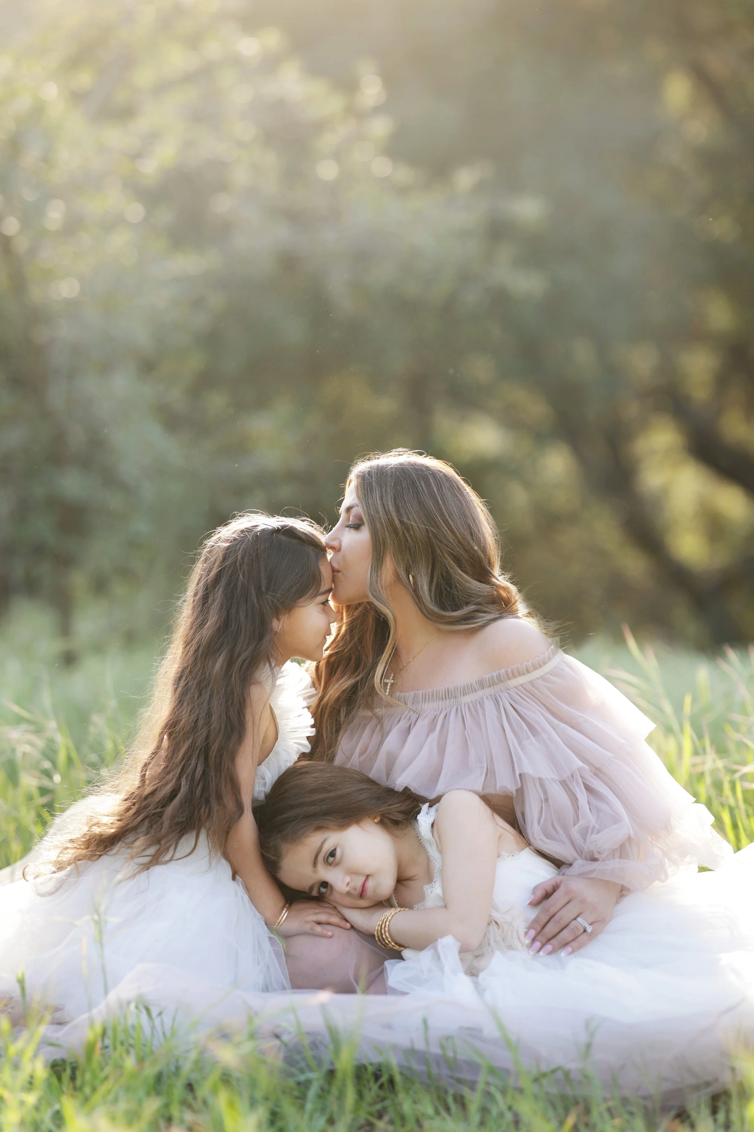 A woman with two young girls sitting on grass in a park during the golden hour, one girl resting her head on the woman's lap, the woman kissing one girl's forehead.