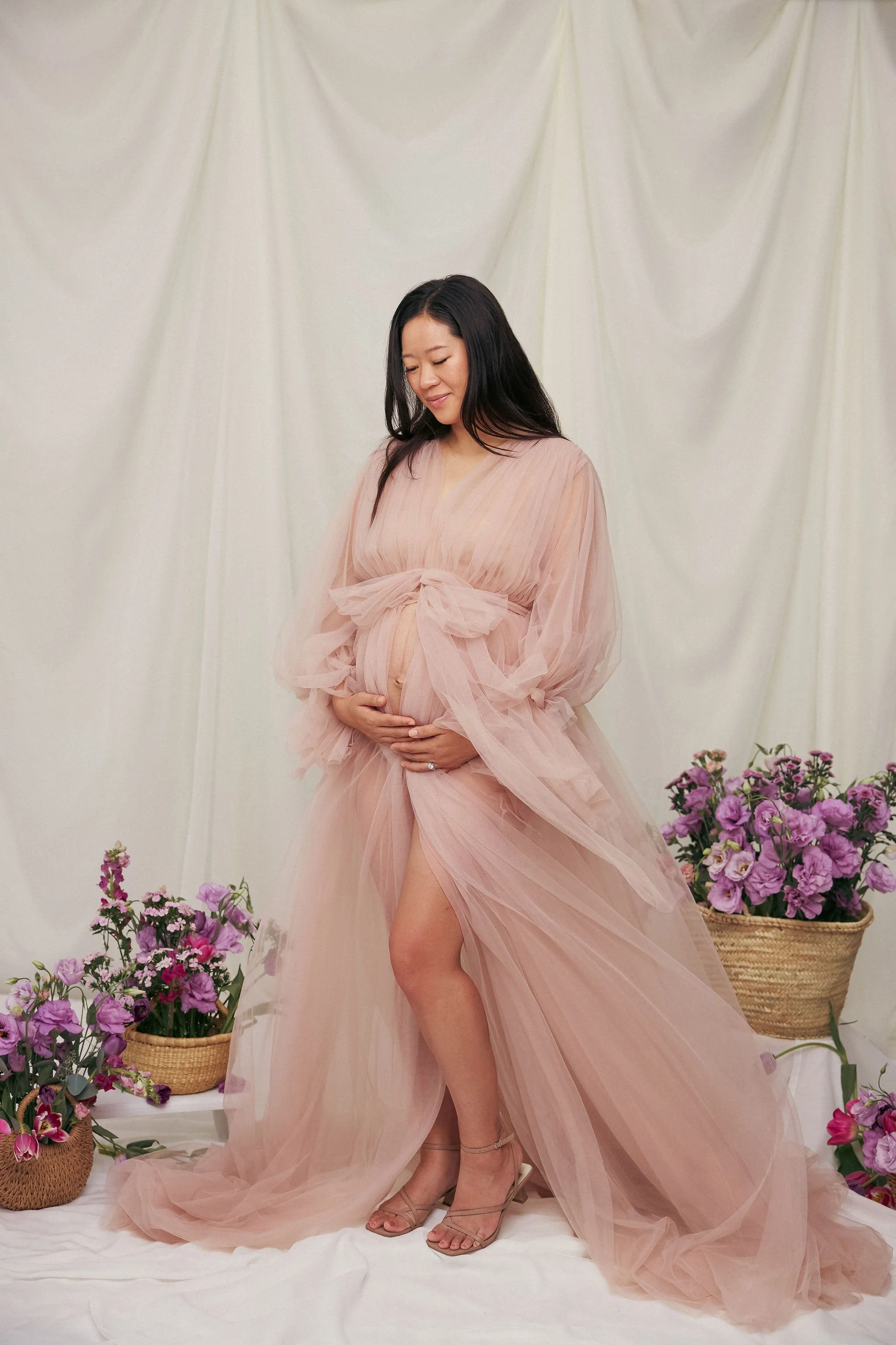 A pregnant woman in a flowing, blush-colored gown with a slit, standing in front of a cream-colored backdrop with baskets of pink and purple flowers around her.