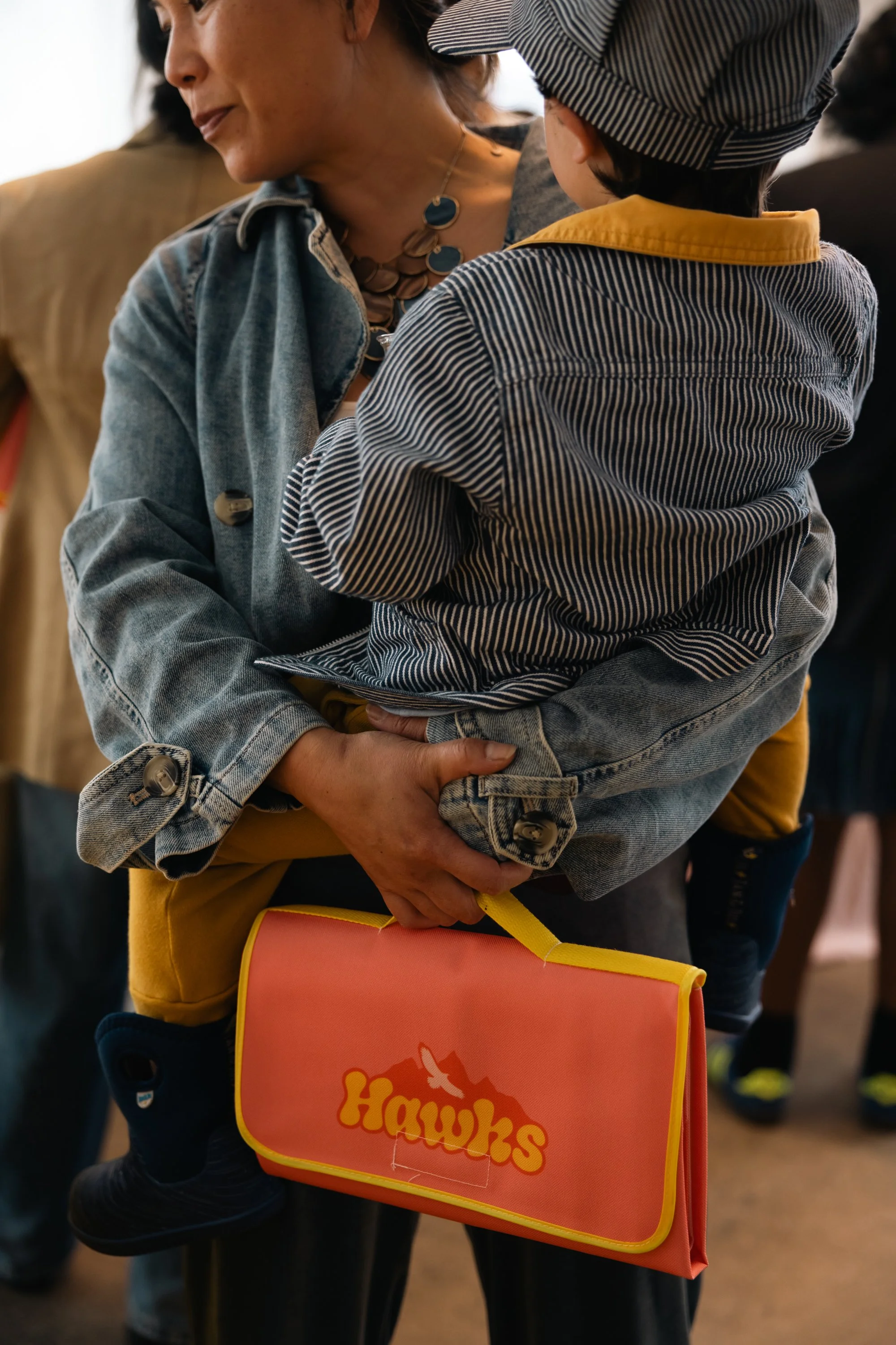 A woman holding a small child who is wearing a striped jacket and a cap. The woman is wearing a denim jacket and a necklace. The woman is carrying a pink bag with the word 'Hawks' and a mountain logo on it.