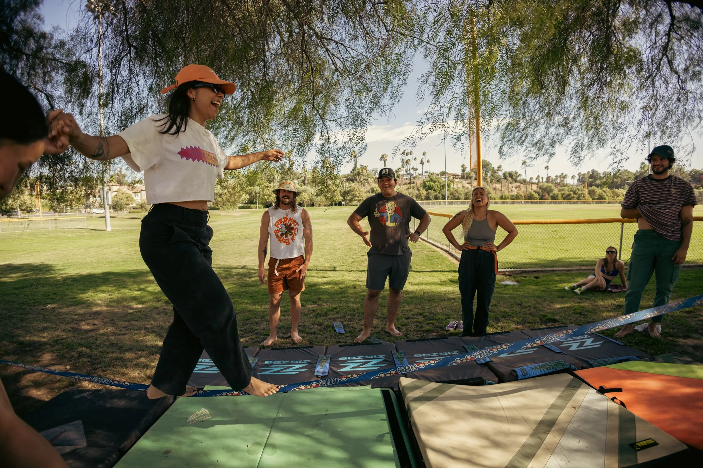 Group of people outdoors under a tree, laughing and playing on a slip and slide in a grassy park area.