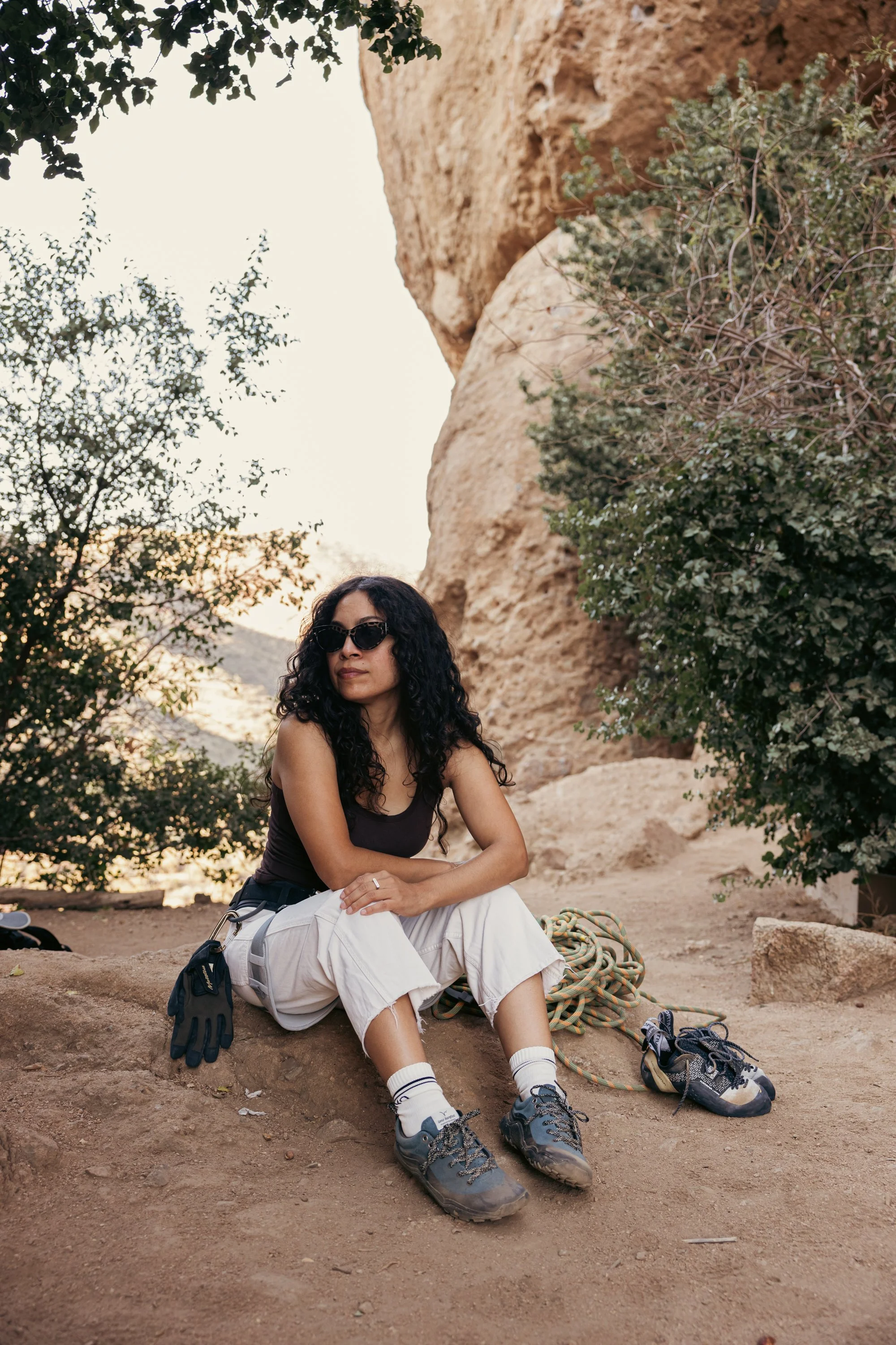 A woman with curly black hair, sunglasses, and a black tank top sitting on a rock in a desert landscape with rock formations and green shrubs.