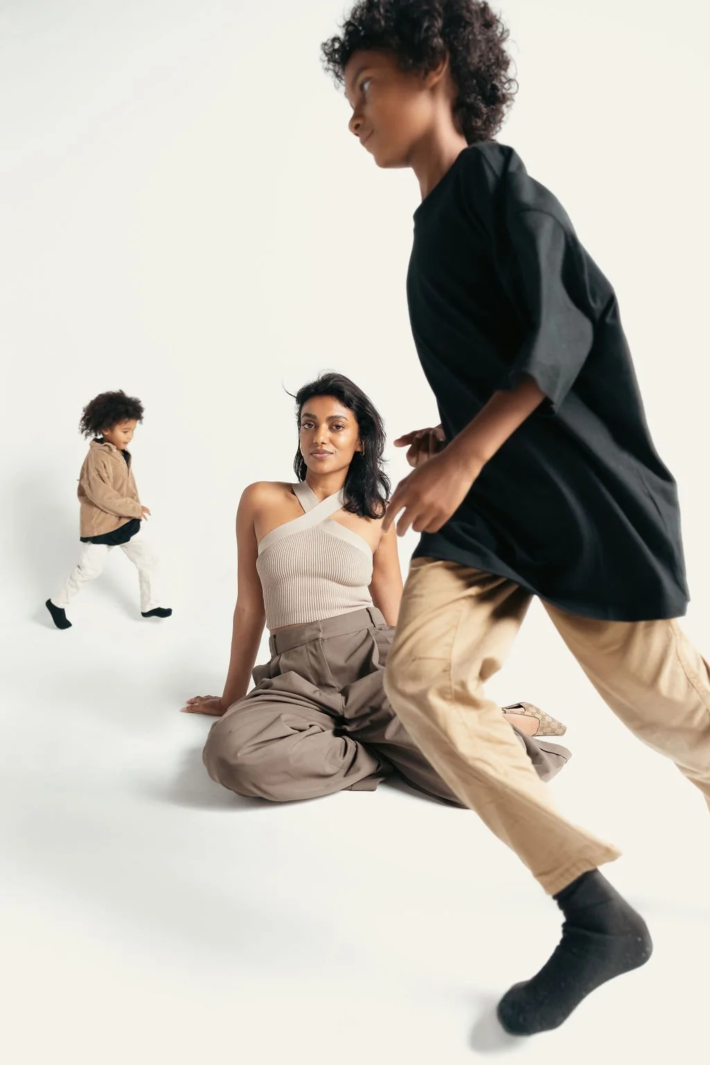 A woman sitting on the floor with two children playing nearby against a plain white background. Fun Family photograph