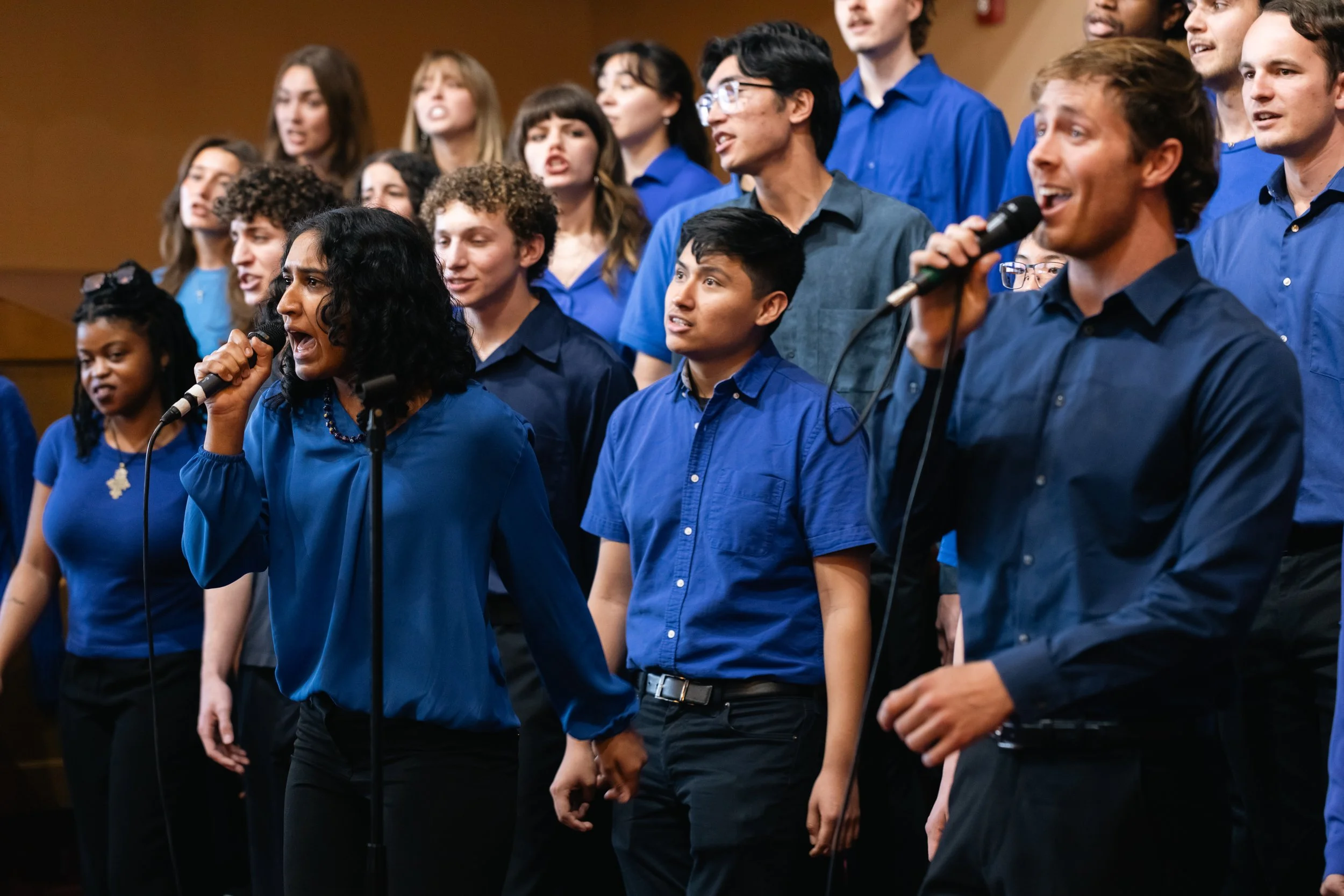 A diverse group of people singing together on stage, wearing blue shirts, with two individuals in the front holding microphones.