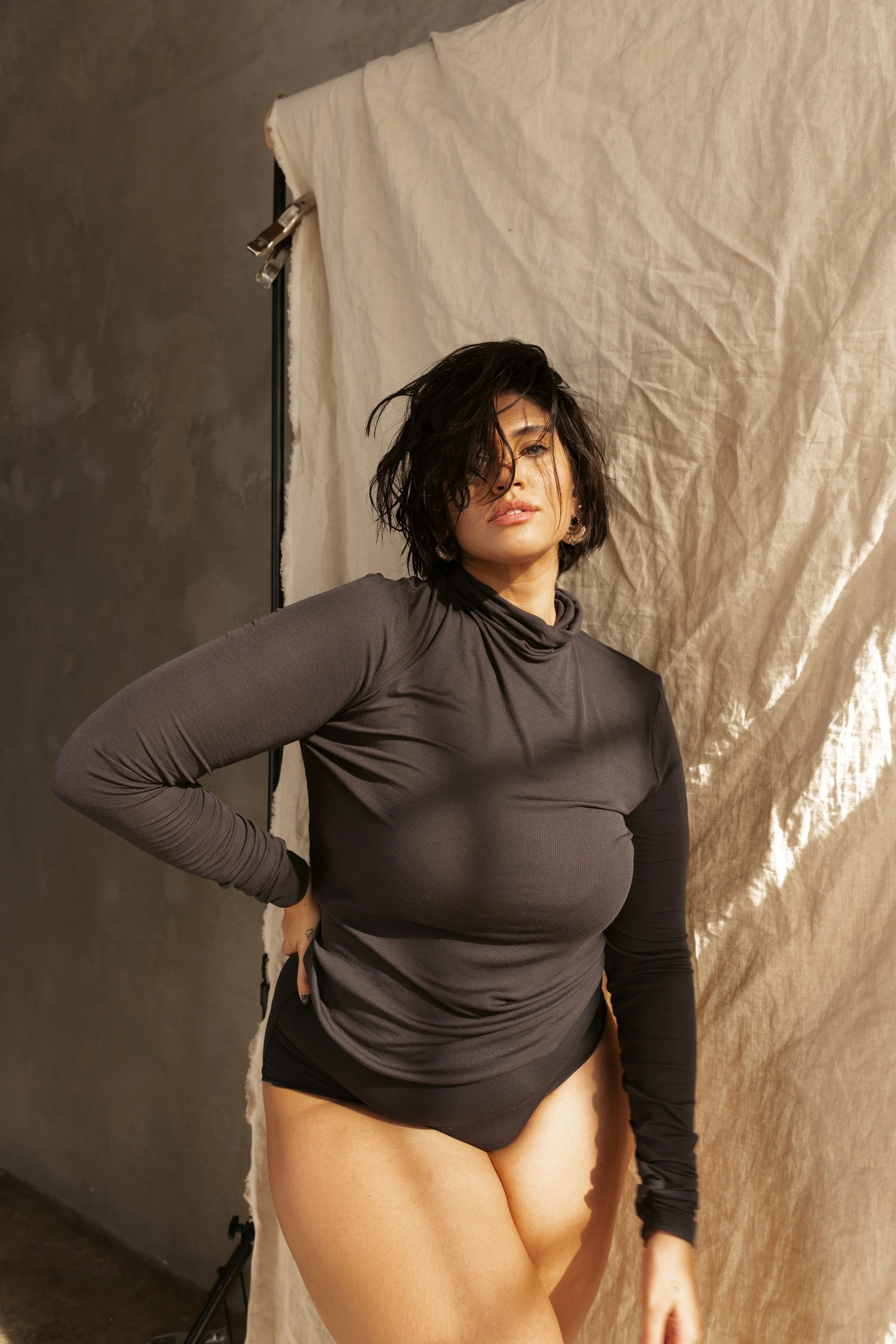 A woman with dark, messy hair and wearing a long-sleeve black top is posing in front of a beige backdrop.