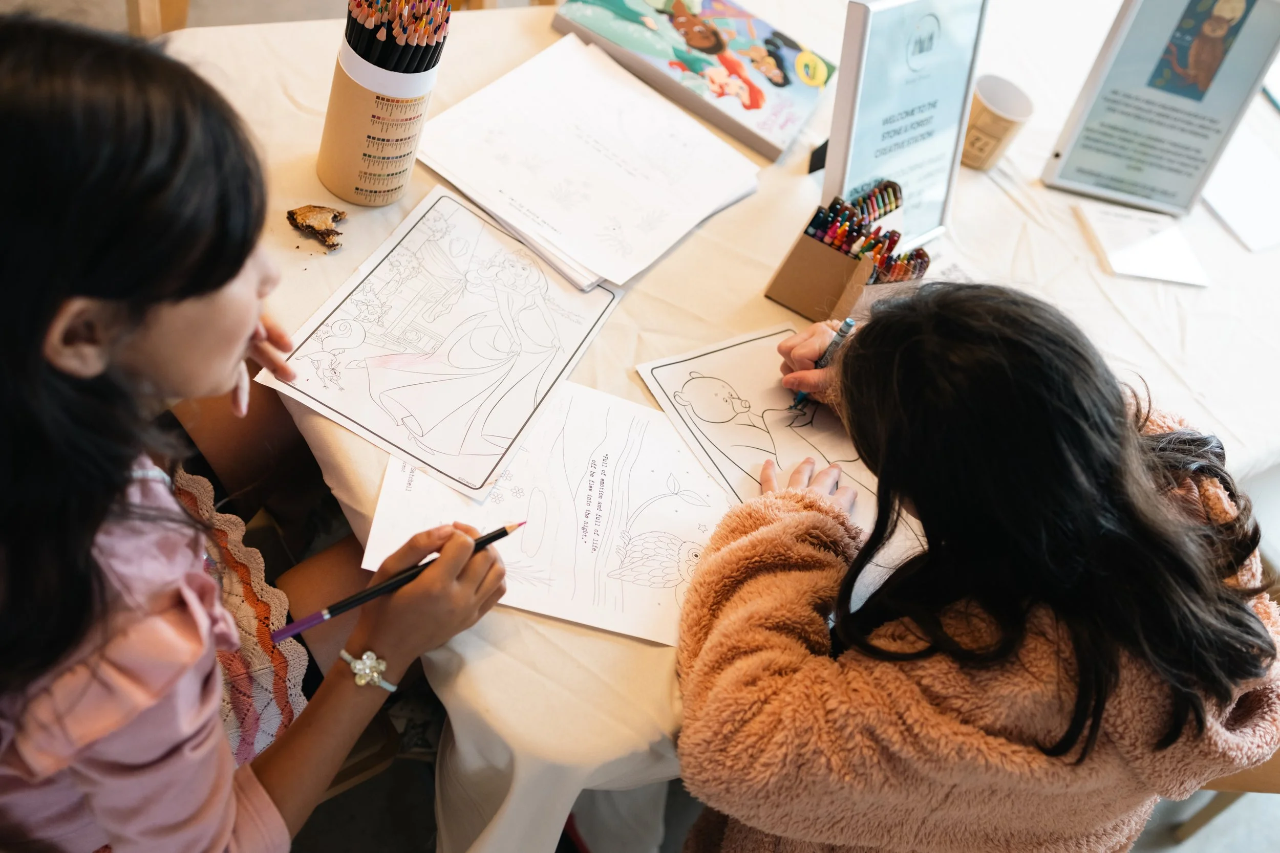Two young girls sitting at a table, coloring and drawing with coloring books, pens, and papers with various illustrations.