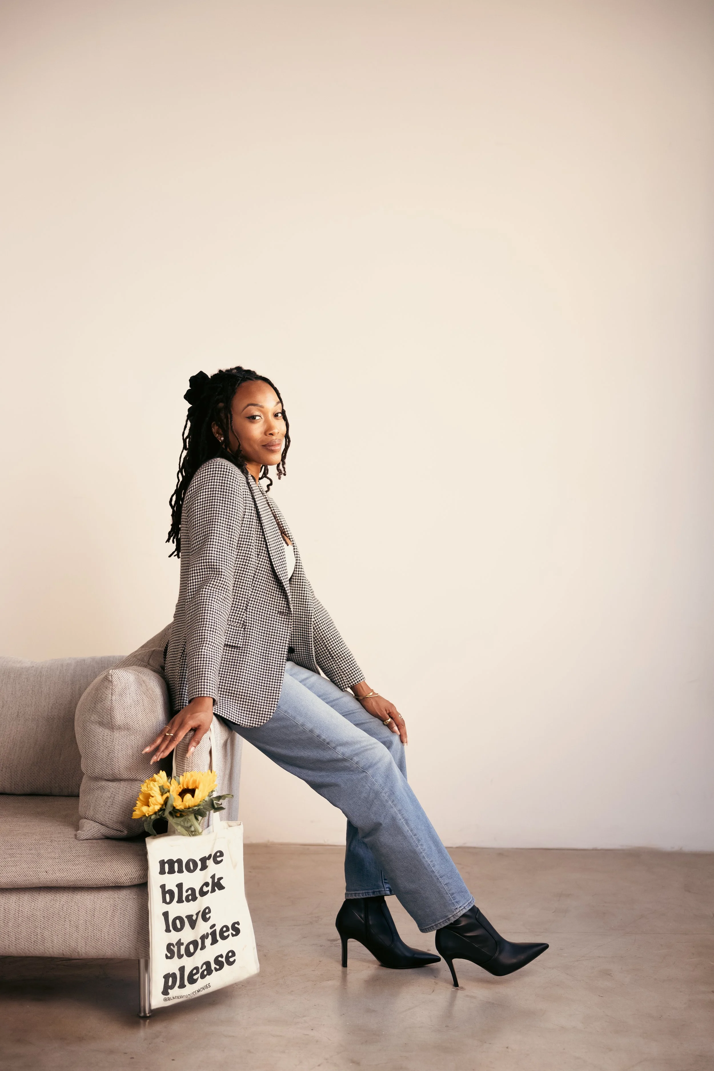 A woman sitting on the edge of a beige sofa, wearing a checkered blazer, light blue jeans, and black high-heeled boots, with a tote bag with sunflowers and the text "more black love stories please" on it, against a plain white wall.