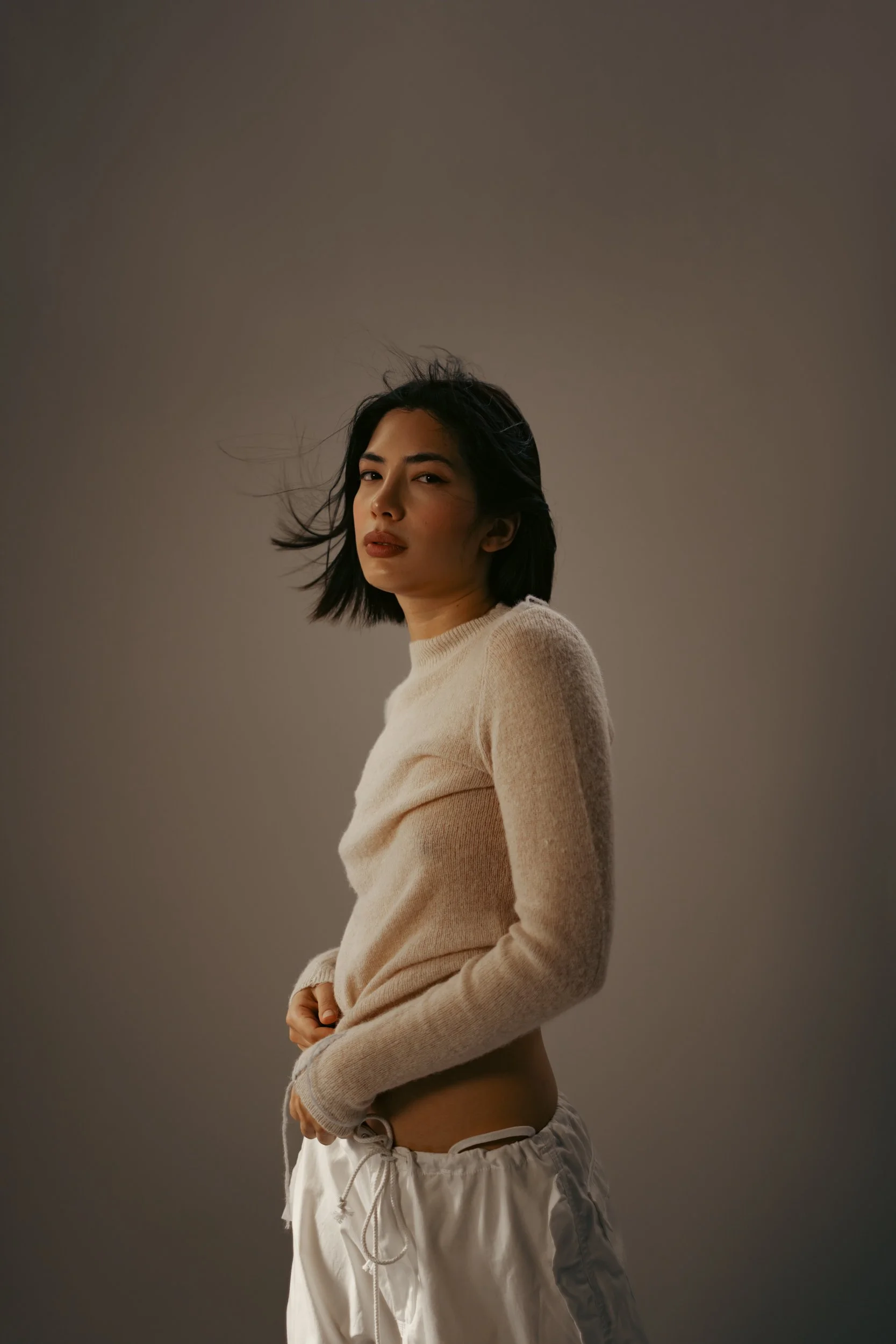 A woman with shoulder-length black hair wearing a beige sweater and white drawstring pants against a plain gray background.