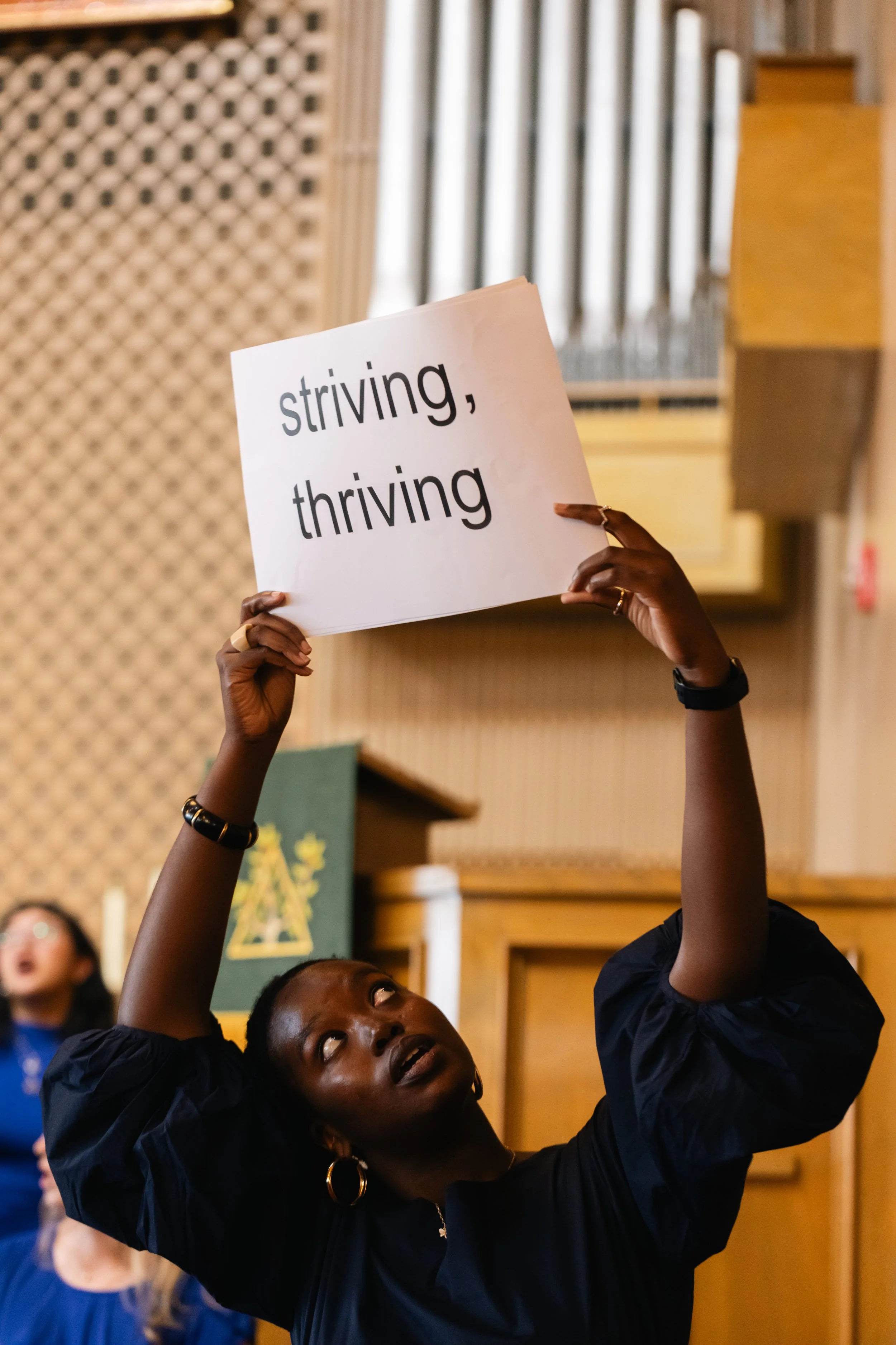 A woman holding a sign that reads 'striving, thriving' inside a room with wooden walls and a large organ in the background.