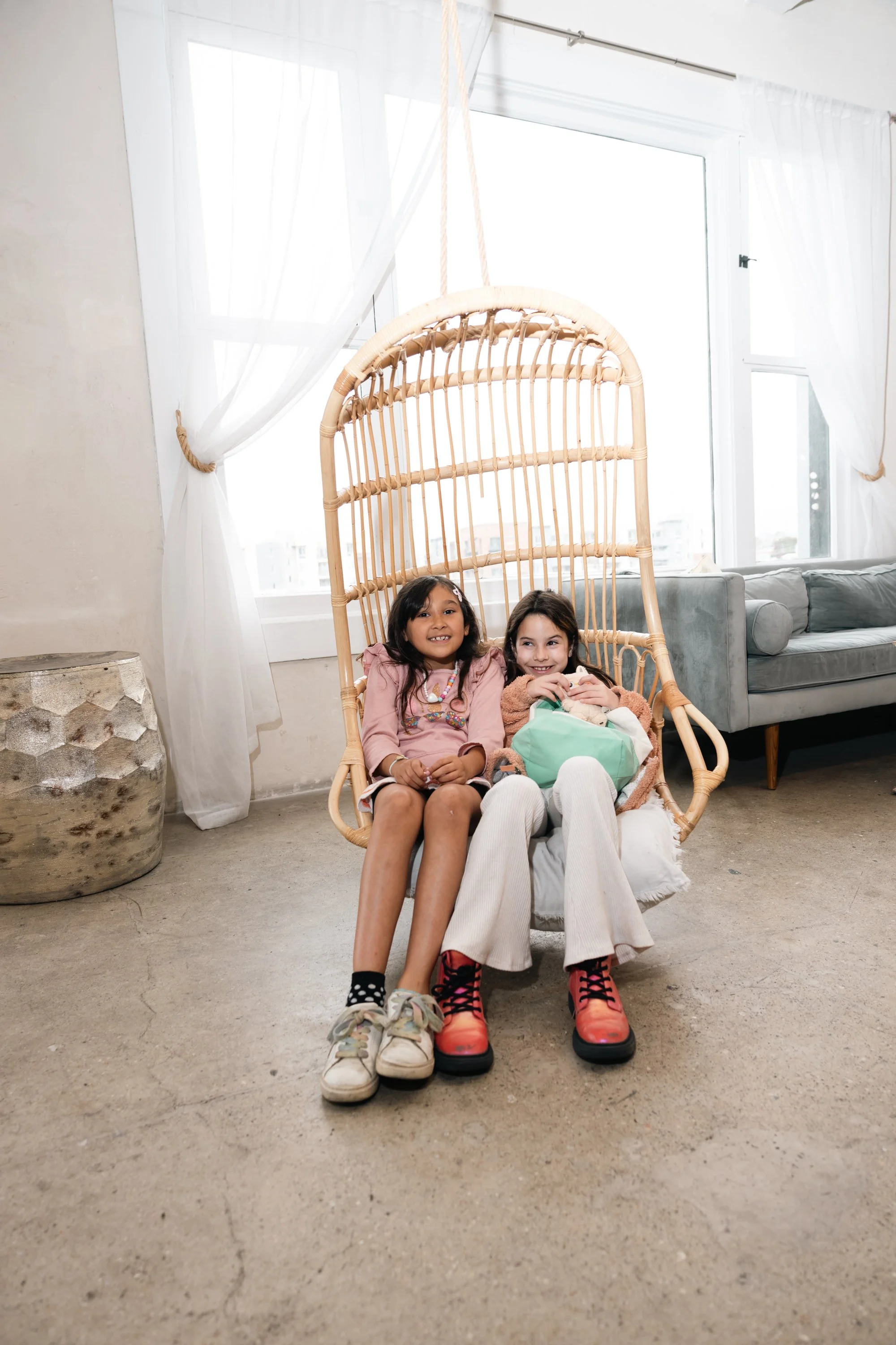 Two young girls sitting closely together in a hanging rattan chair in a bright room with large windows and white curtains. The girl on the left is wearing a light pink top and black shorts, and the girl on the right is wearing a tan sweater and white
