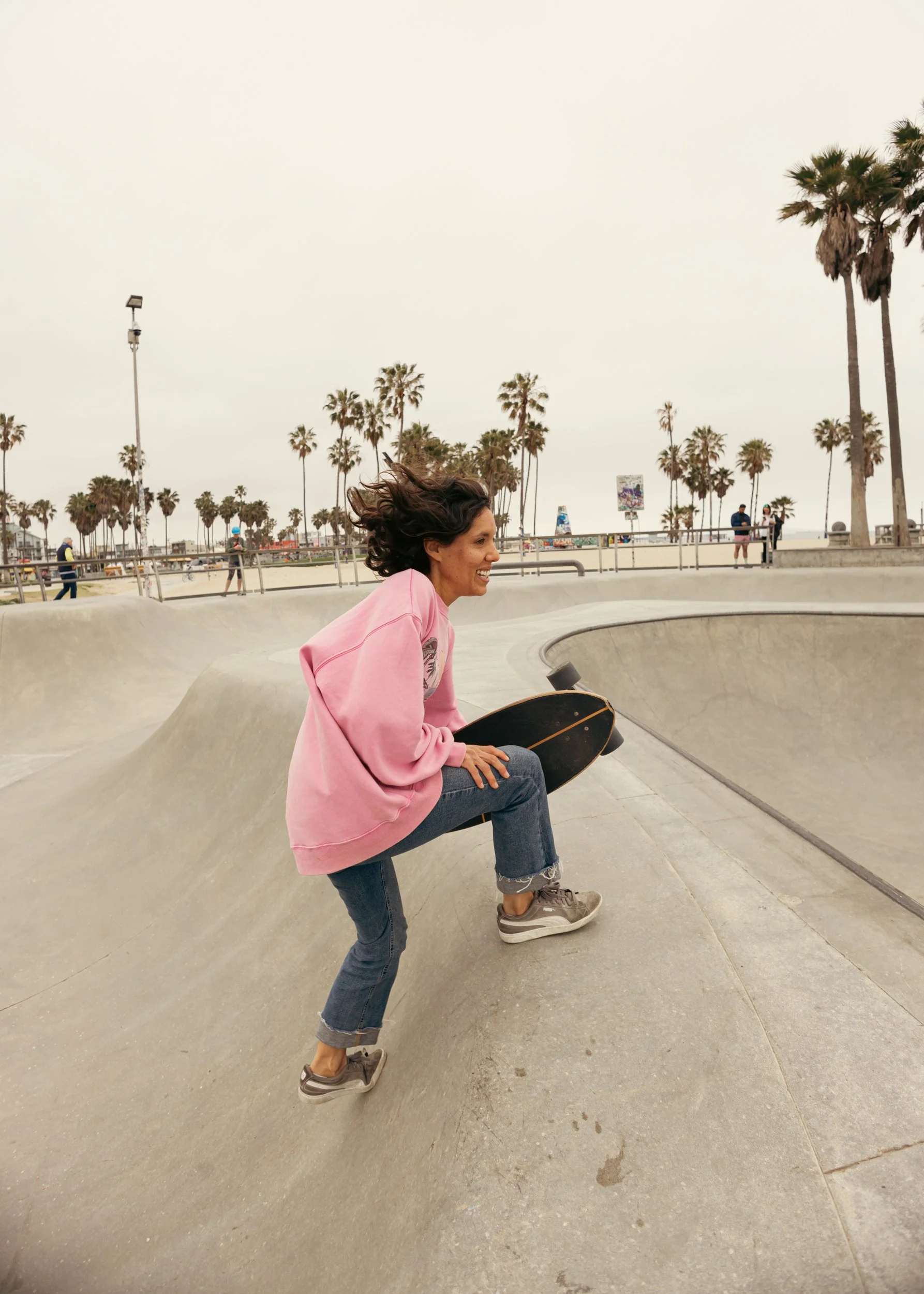 A woman in a pink sweatshirt and jeans skateboarding at a skate park on a cloudy day, with palm trees in the background.