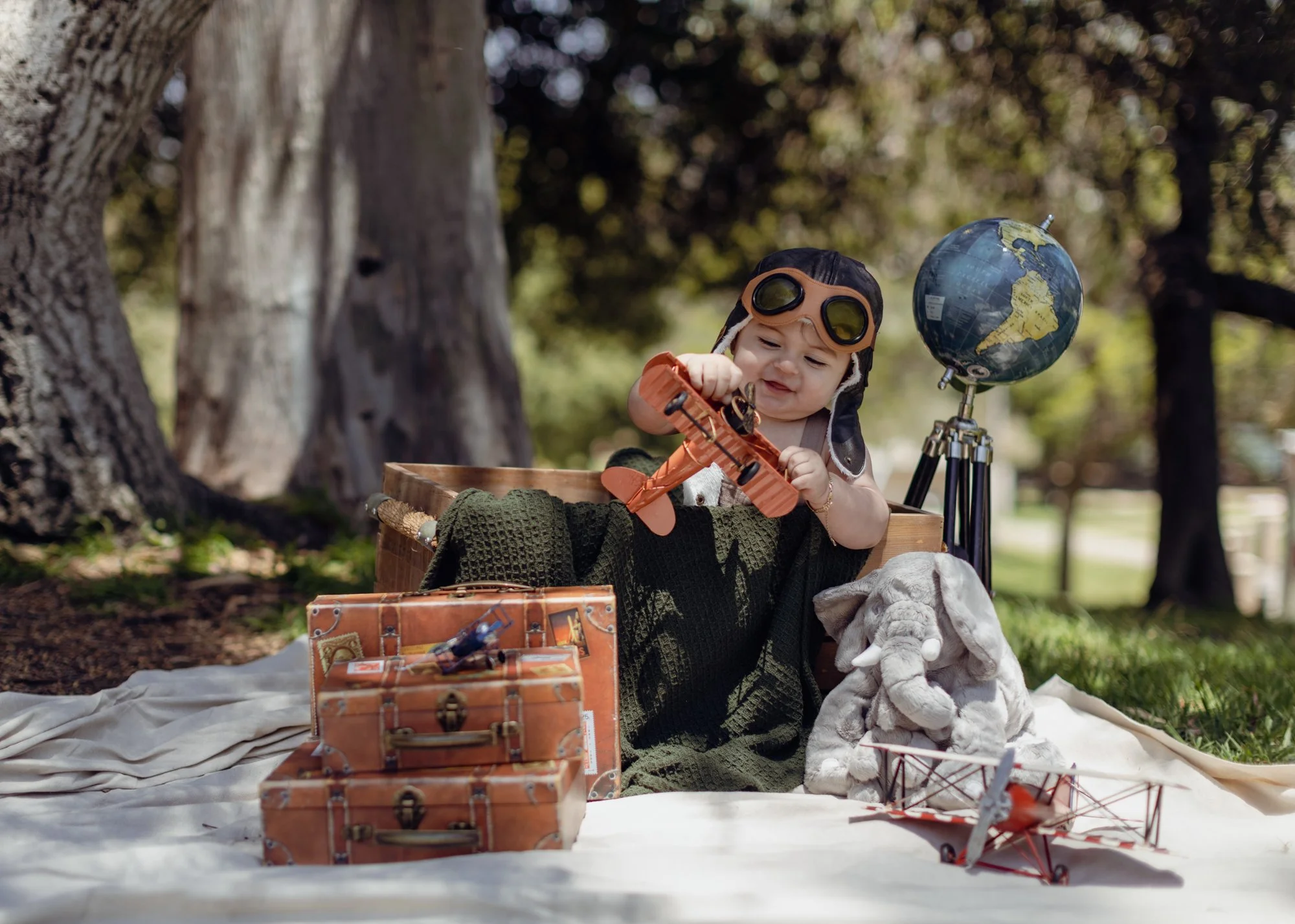 A young child dressed as an aviator playing outdoors with a toy airplane, surrounded by vintage suitcases, a globe, and plush elephant toys, sitting on a blanket in a park under large trees. Outdoor first year birthday photography.
