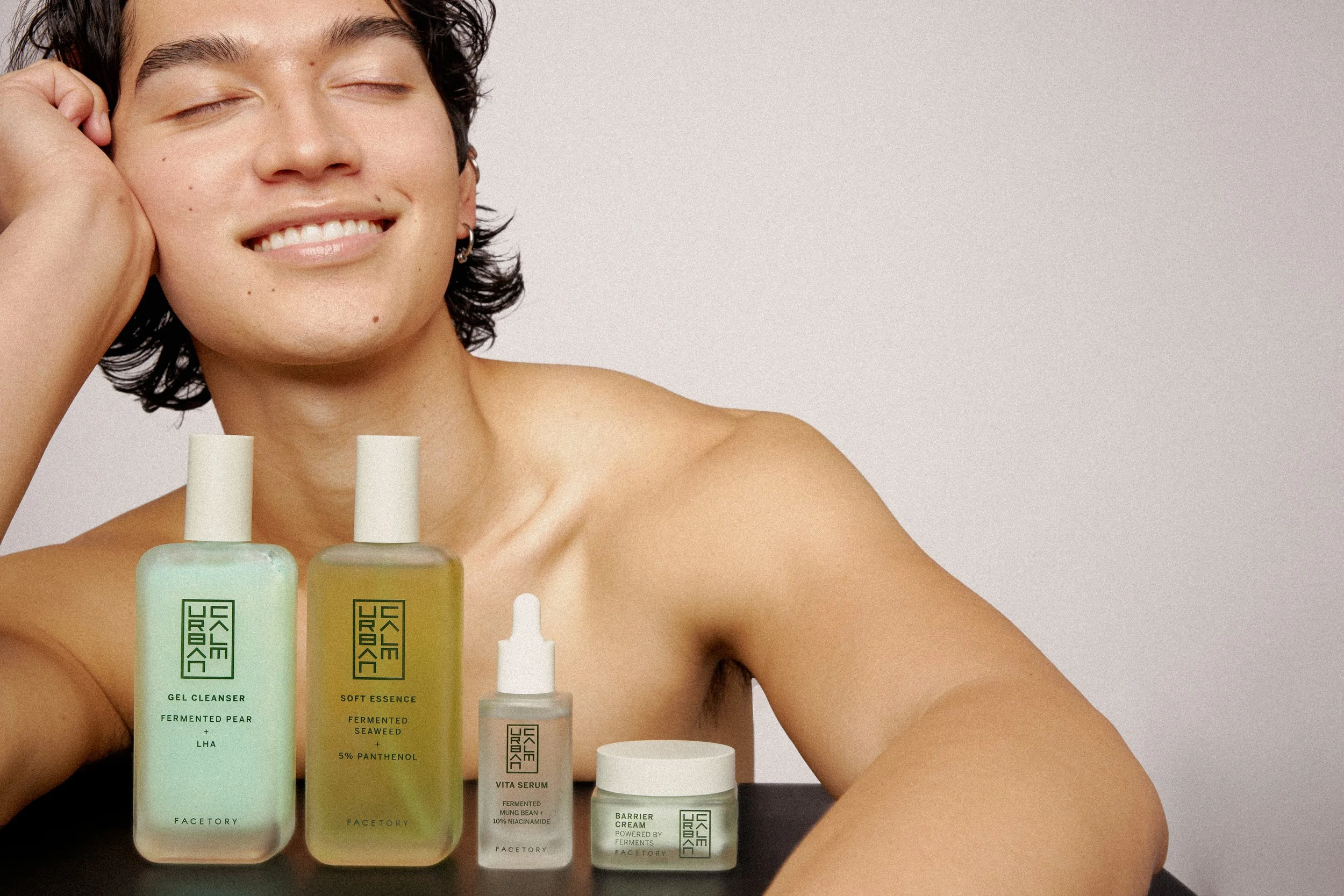 A smiling man with short dark hair has her eyes closed and rests his head on his hand. In front of her, there are five skincare products including gel cleanser, serum, moisturizer, and creams on a black surface against a plain light background.