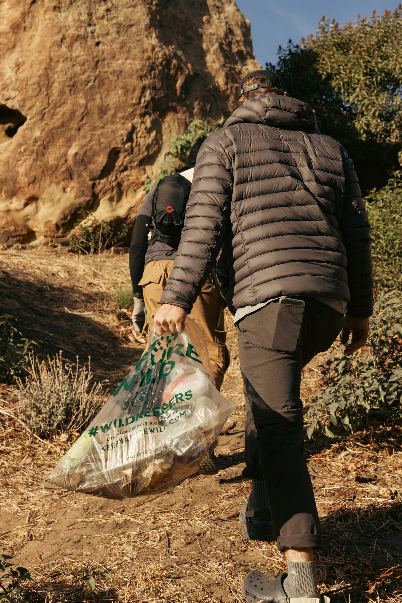 People hiking on a trail, one holding a large plastic trash bag.