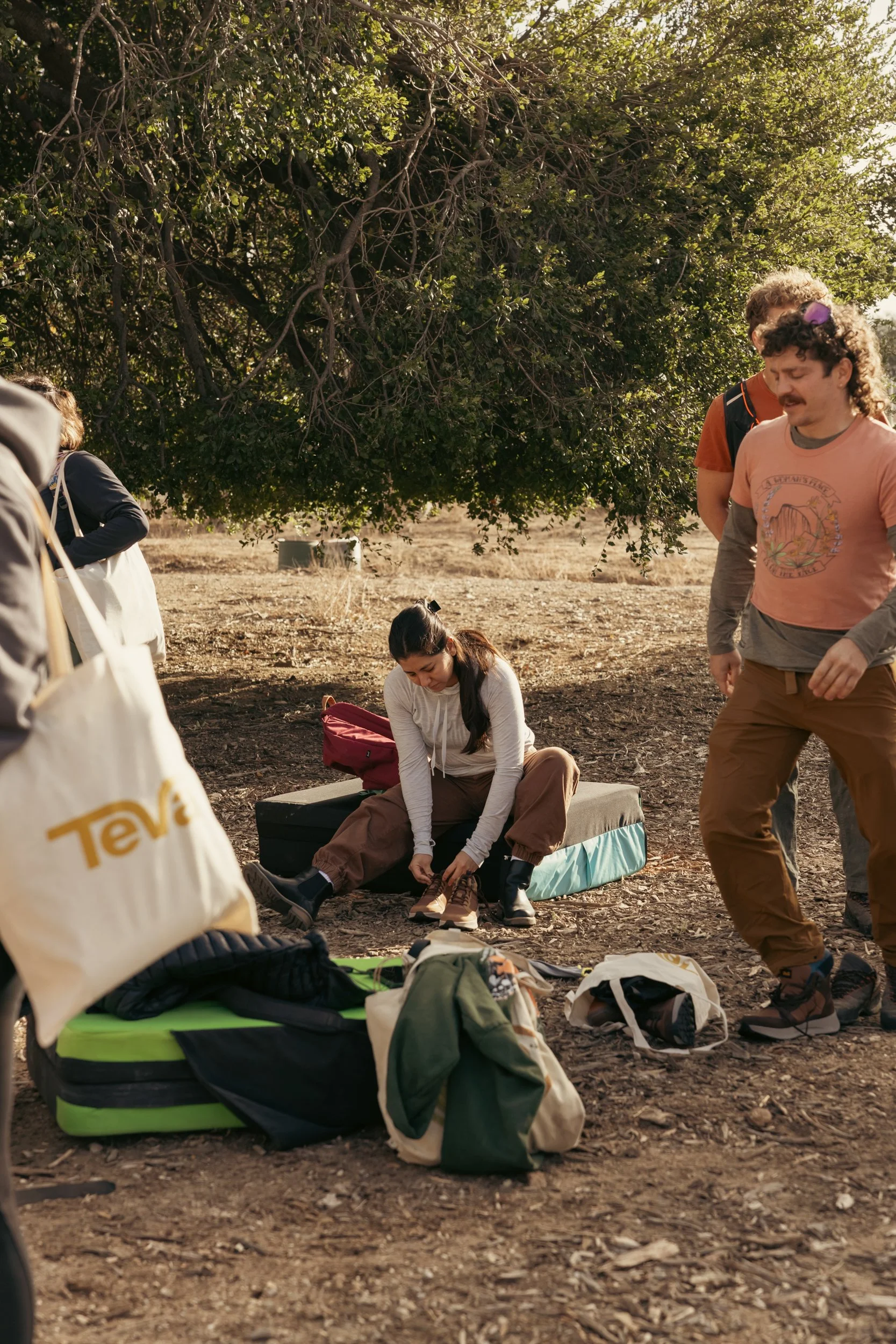 Group of people outdoors preparing for hiking, with backpacks, sleeping pads, and other gear on the ground, under a large tree.