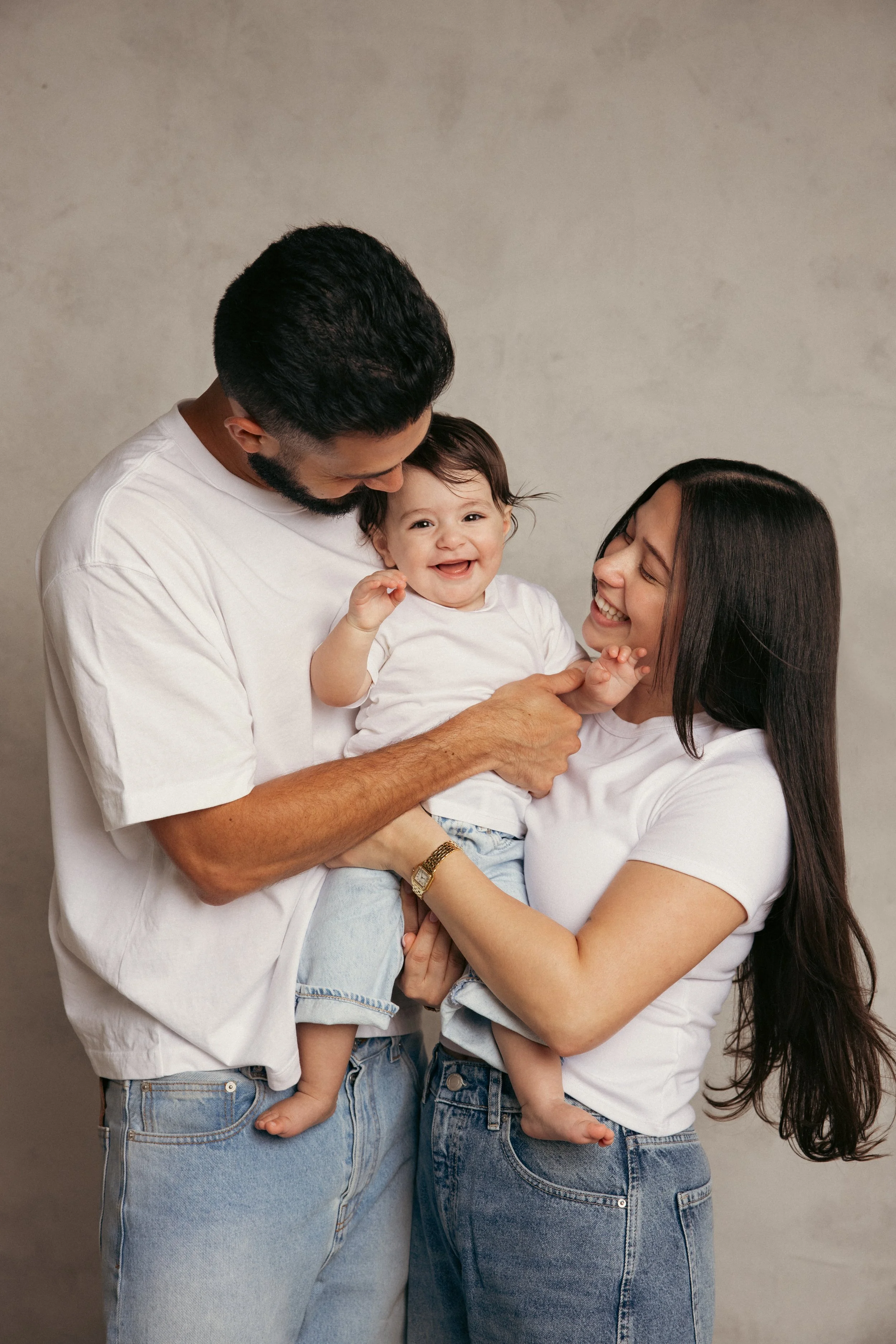 A happy family of three, with a father, mother, and toddler, smiling and playing together indoors.