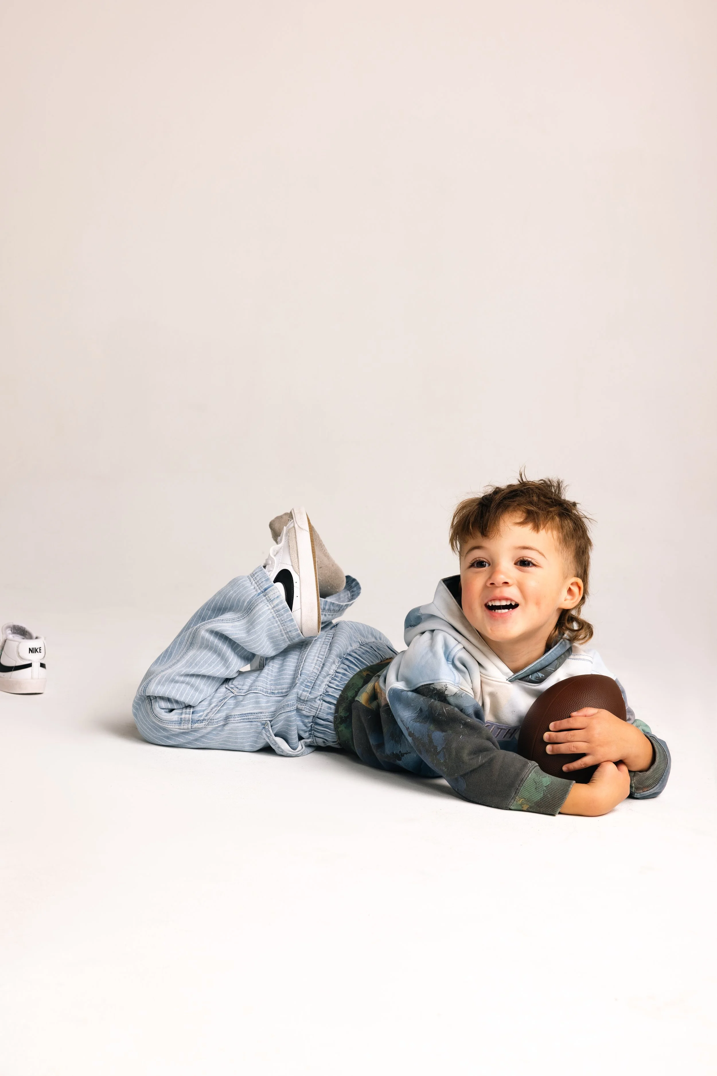 Young boy lying on the floor holding a football, smiling, with a pair of Nike sneakers nearby on a plain white background.
