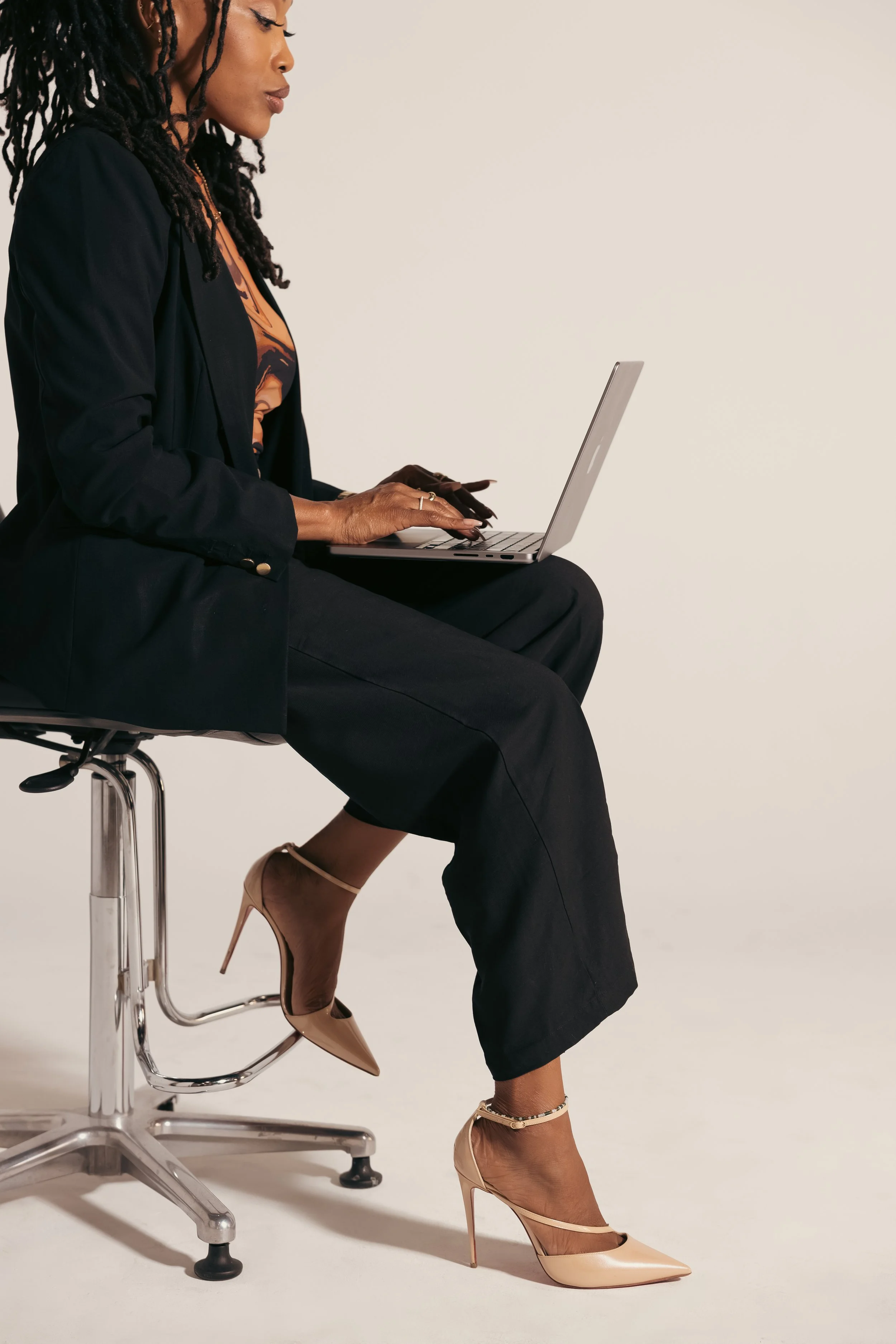 A woman with long braids is sitting on an office chair, using a laptop. She is wearing black pants, a black blazer, nude high heels, and an orange and black patterned top.