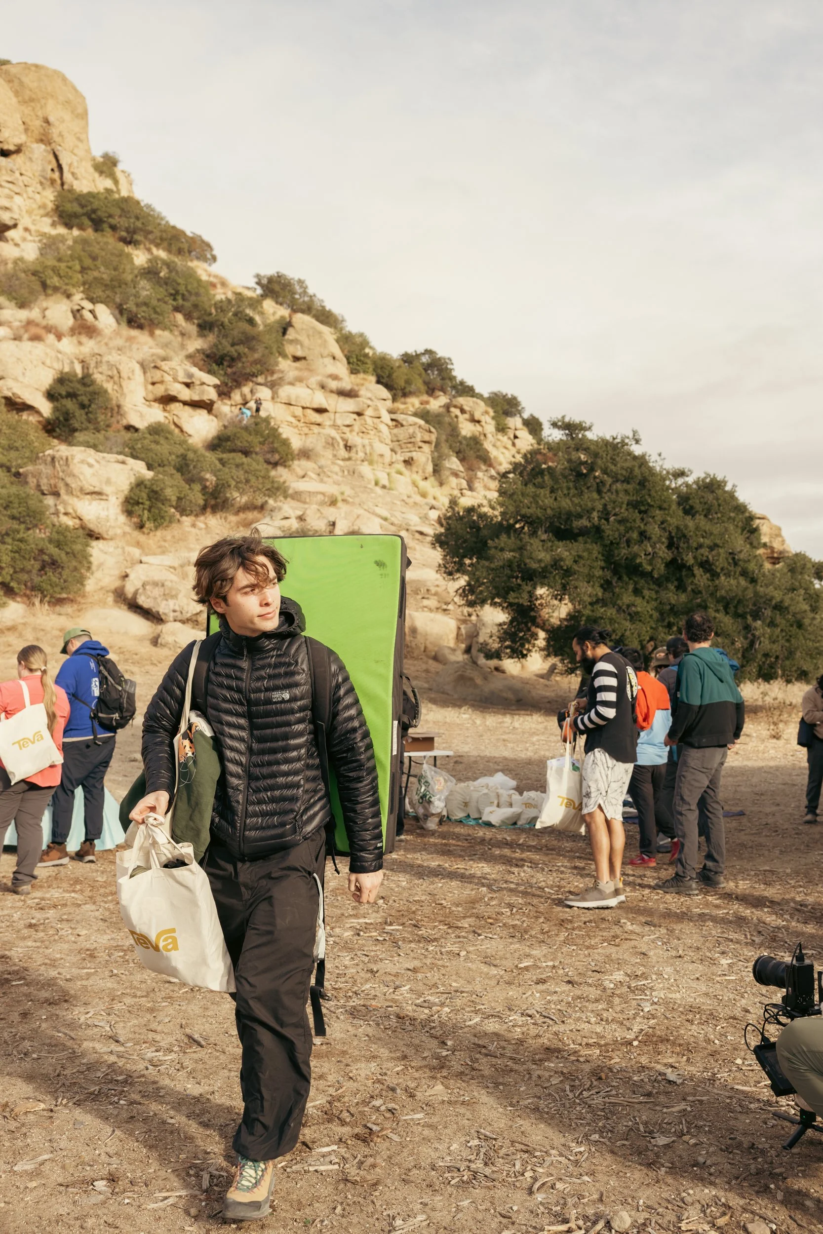 Young man with a large green climbing pad on his back, carrying a reusable bag, walking outdoors with a group of people in a rocky, rural area.