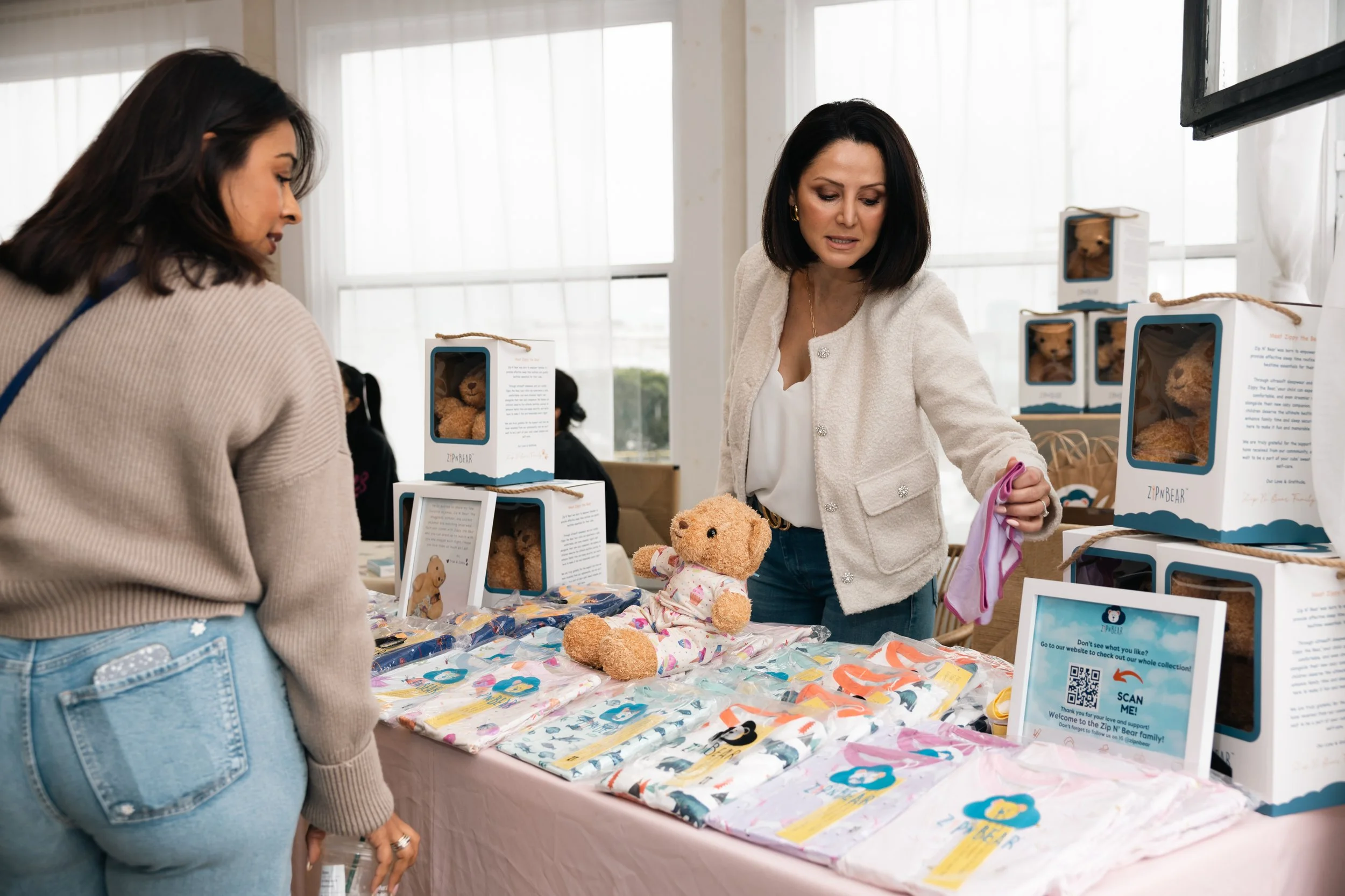 Two women browsing a table of teddy bears and teddy bear-themed merchandise at an indoor market