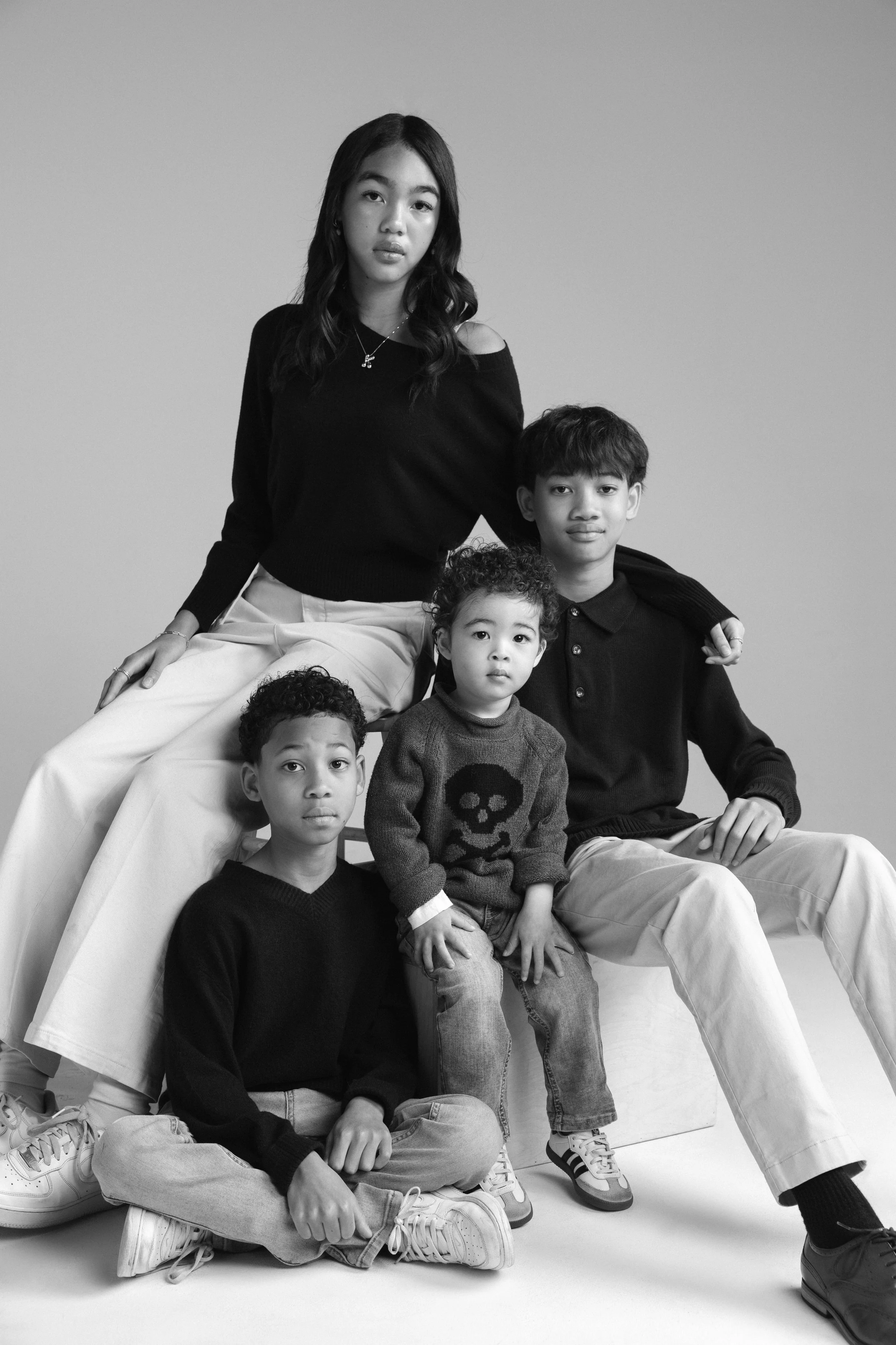 A black and white photo of five children posing together against a plain background. One girl is sitting on a high stool, with a boy standing behind her, and three other children sitting on the ground in front.