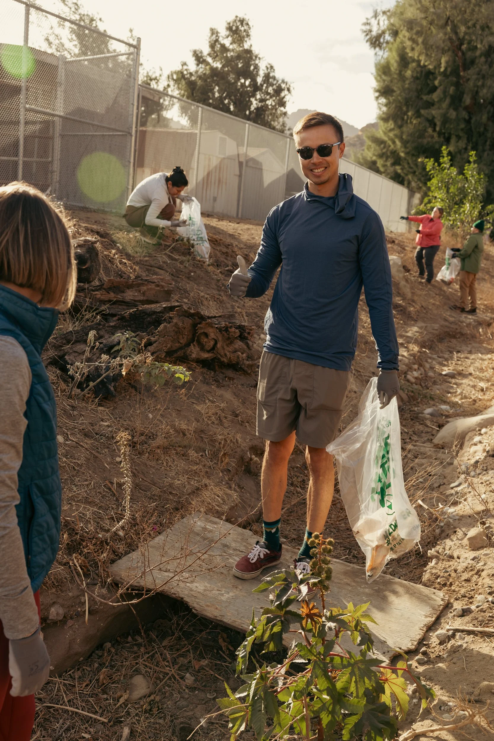 Group of people participating in a community cleanup outdoors on a sunny day, with trees and a fence in the background. A man in the foreground is smiling, wearing sunglasses, a blue long-sleeve shirt, tan shorts, and holding a trash bag, giving a th