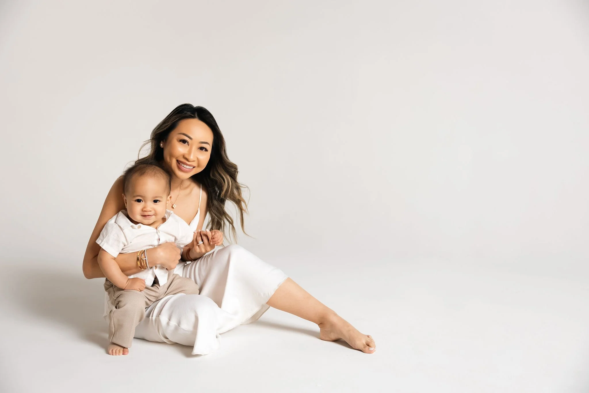 A smiling woman with long wavy hair sitting on the floor with a toddler boy, holding his hand, both dressed in light-colored clothing, in a plain white background.