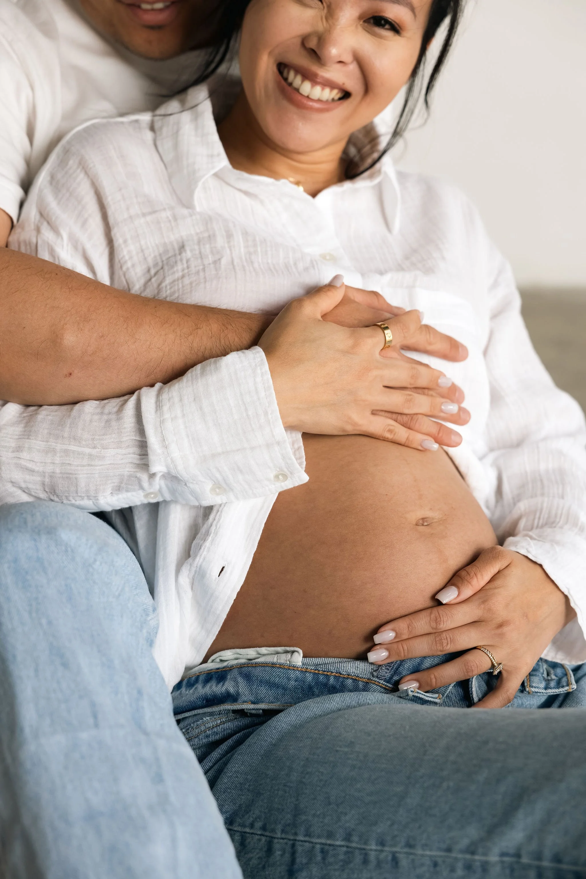 Close-up of a pregnant woman with a joyful expression, wearing a white shirt and jeans, with hands and an adult's arm on her belly, who is also smiling. The adult's hand is on her shoulder, with their arm wrapped around her, showing support.
