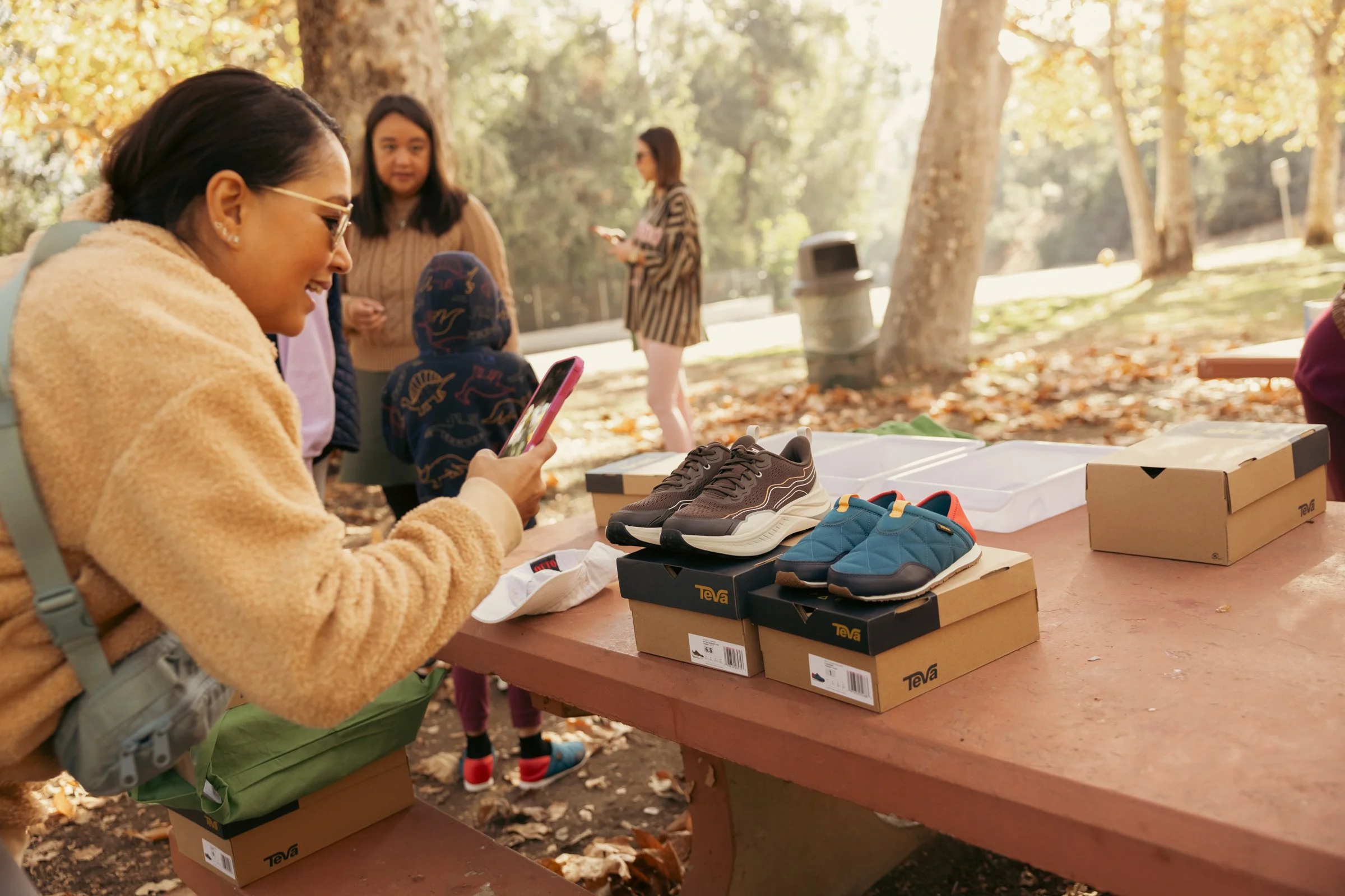 People gather at a picnic table outdoors, looking at and taking photos of new sneakers on display. The table has several boxed shoes, surrounded by trees with autumn leaves.