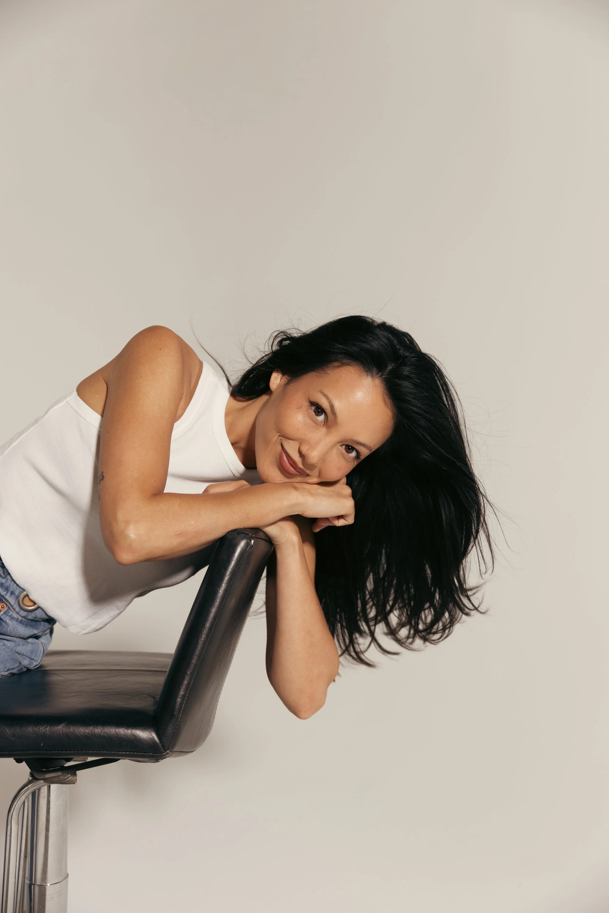 A woman with dark hair leaning on a black office chair, smiling and resting her chin on her hands, against a plain light-colored background.