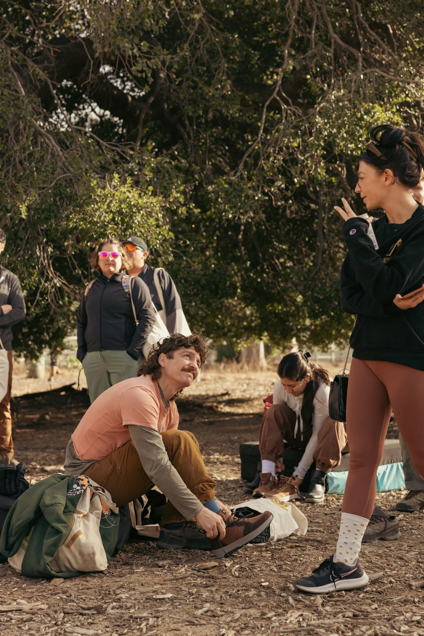 A group of people gathered outdoors in a wooded area, with one man sitting on the ground tying his shoes and others standing or sitting nearby, some wearing sunglasses and carrying backpacks.